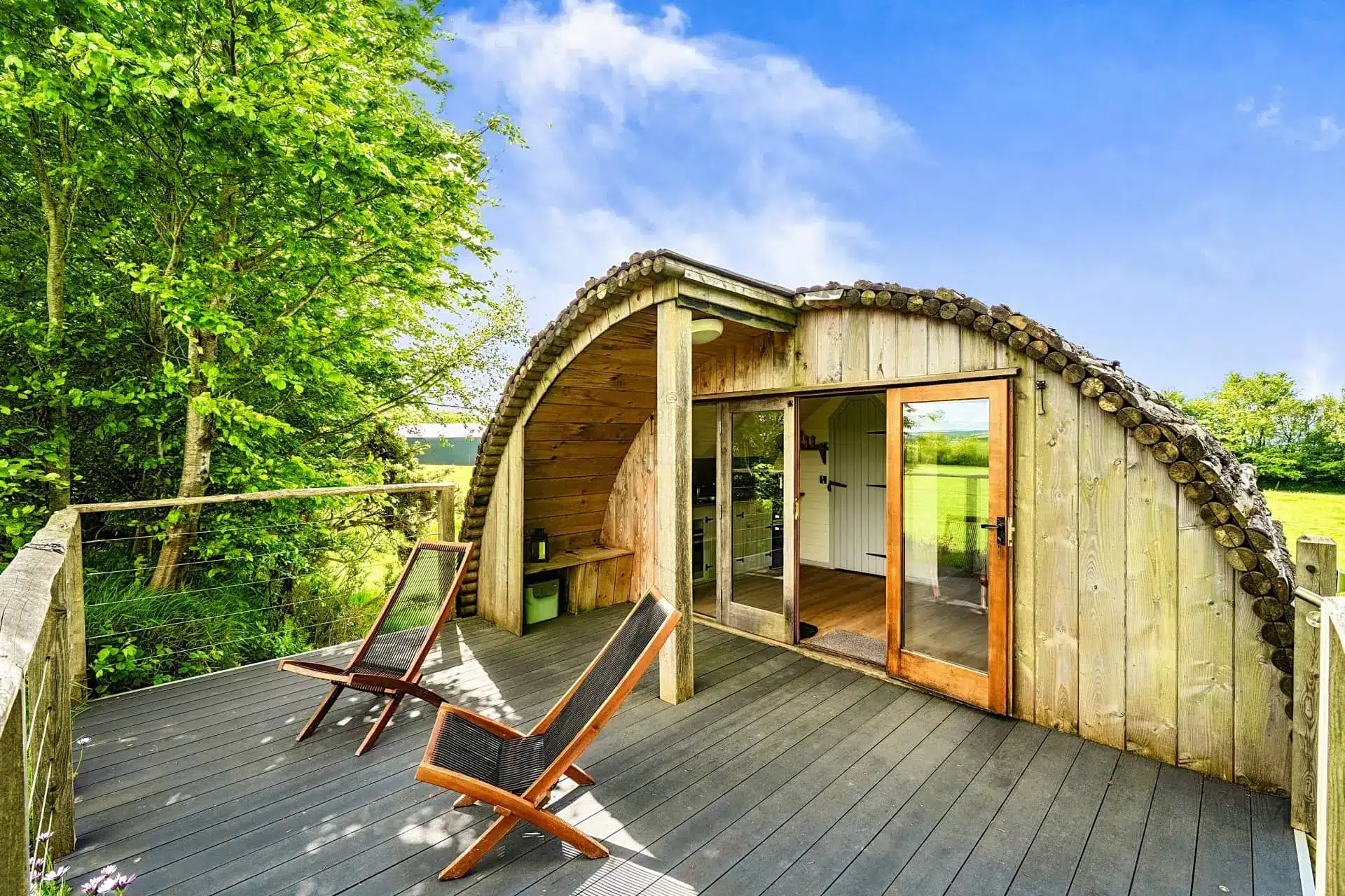 Luxury curved wooden cabin with outdoor deck seating and sliding doors in a green woodland setting