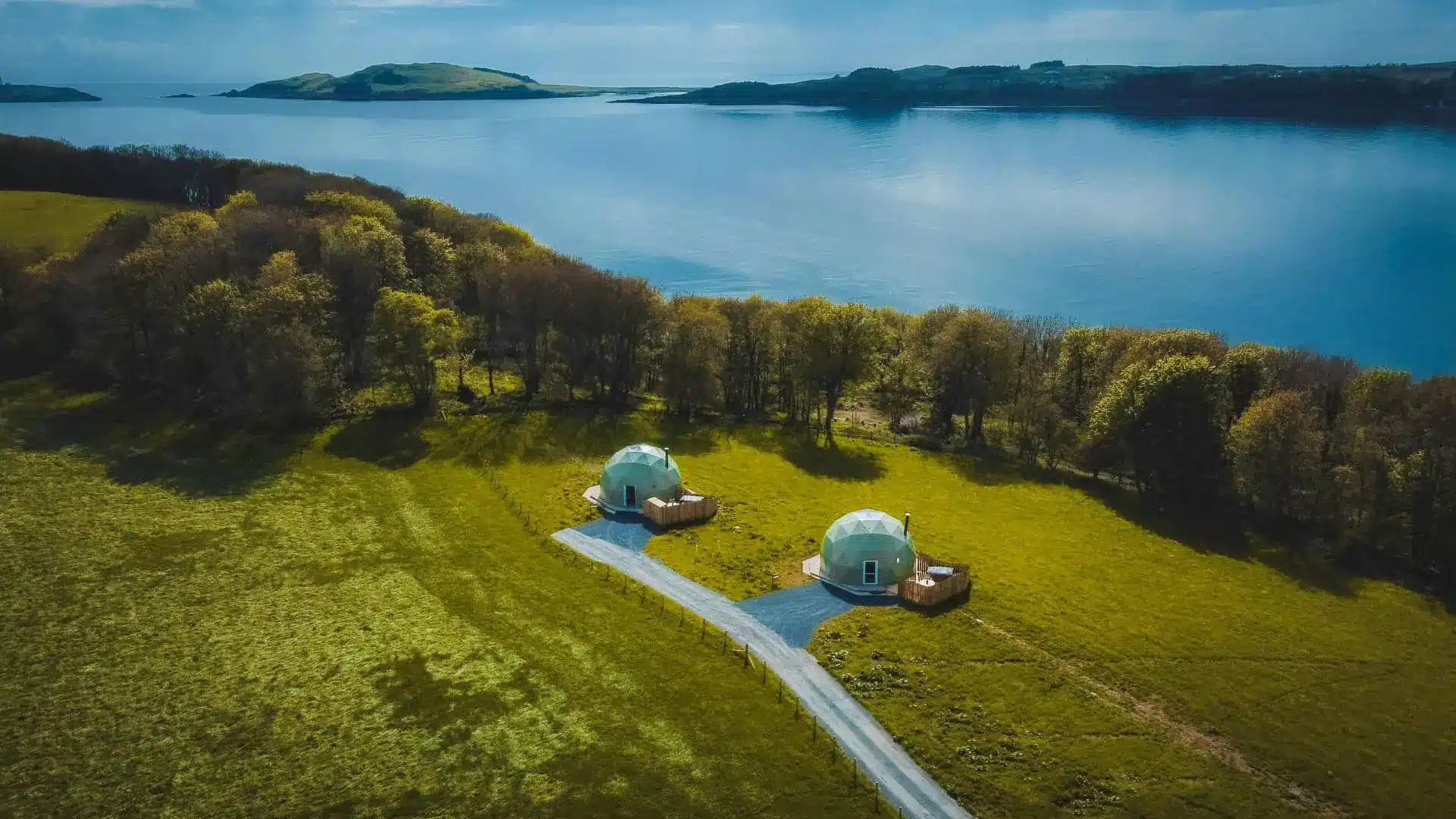 Aerial view of two luxury domed cabins beside a calm lake and lush green grounds