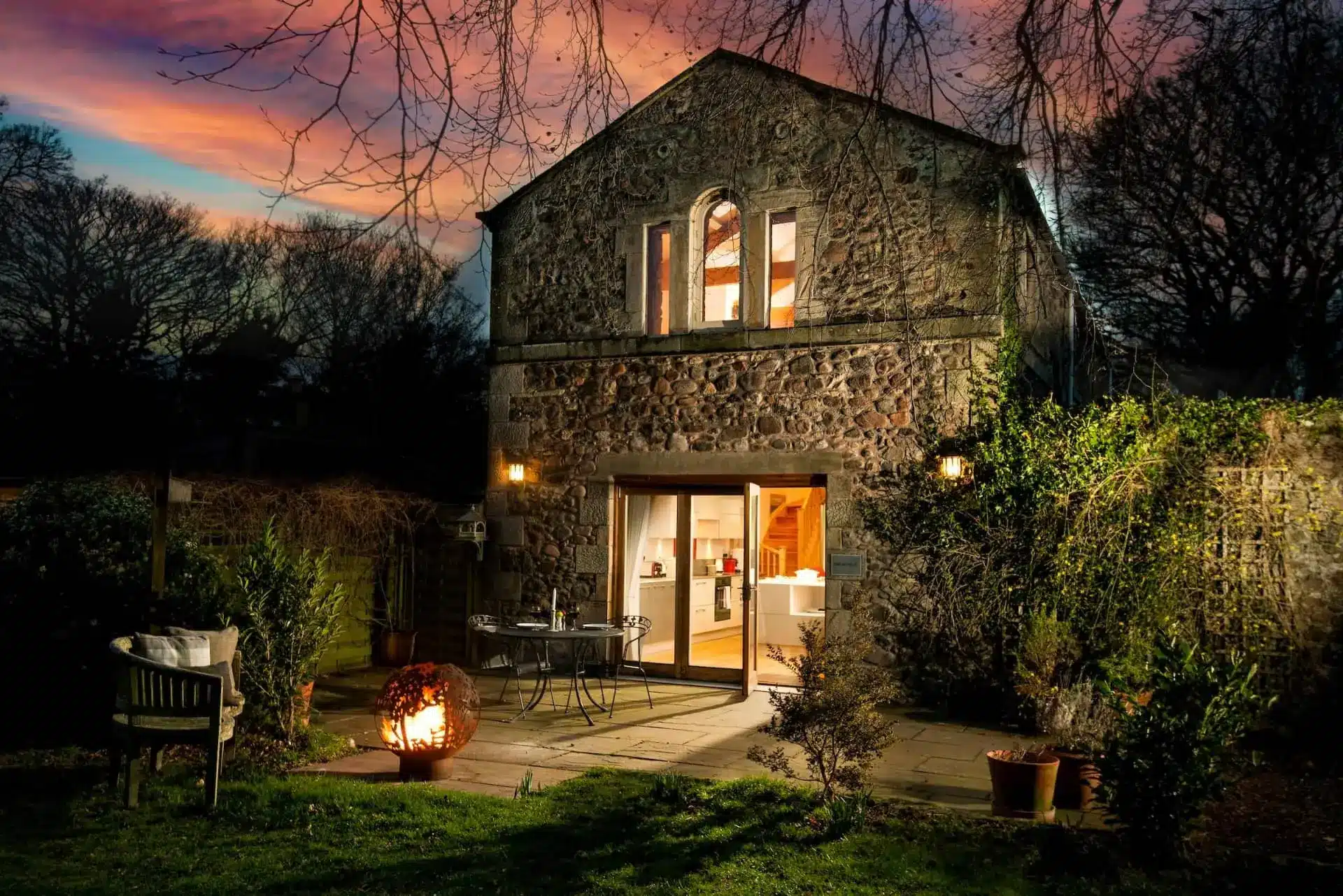 Candlelit evening view of a stone countryside house with open door, patio seating, and glowing lanterns.