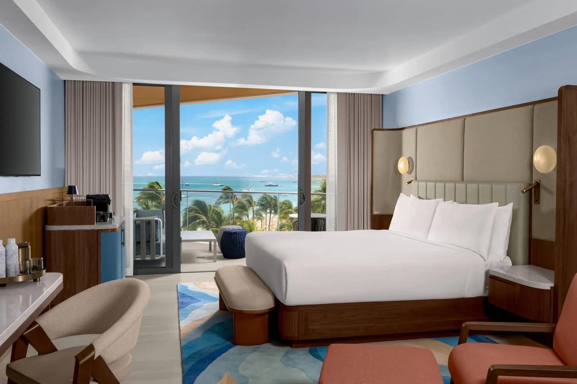 Luxury hotel suite bedroom with a large bed, blue accents, and a view toward palm-lined ocean scenery