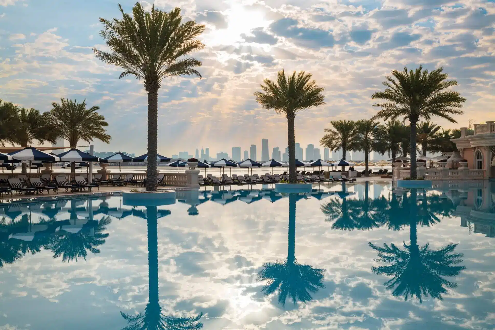 Luxury resort pool lined with palm trees, sun loungers, and a city skyline beyond