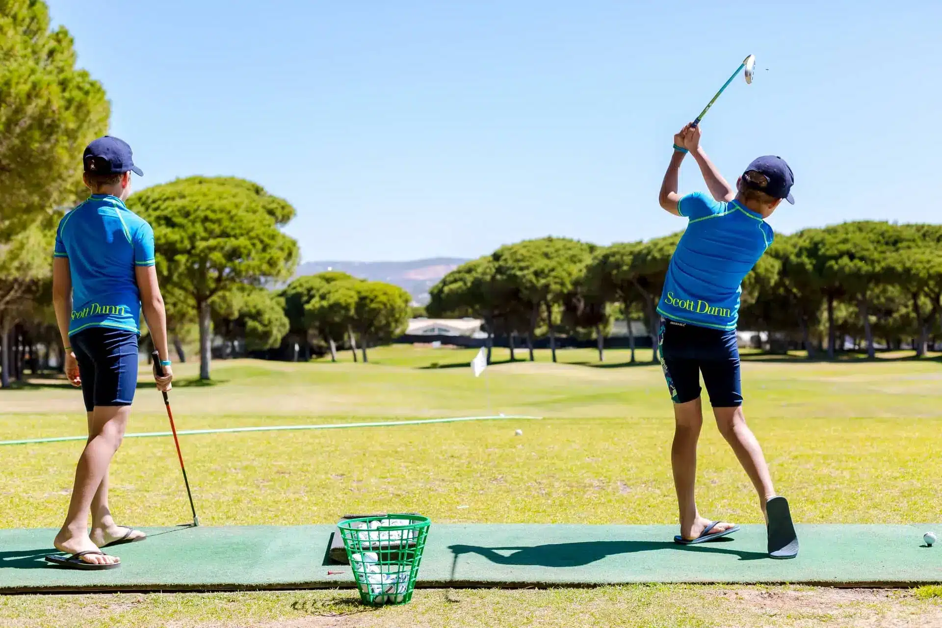 Two young golfers practice on a sunny driving range, one swinging a club beside a basket of balls.