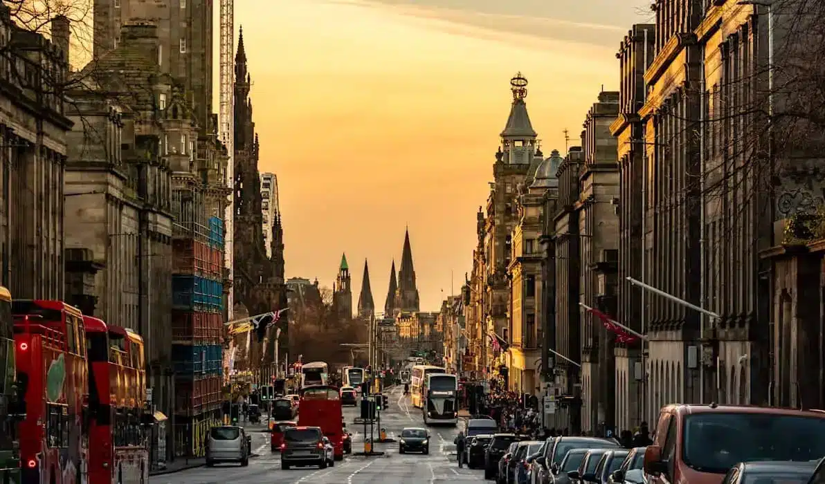 Historic Edinburgh street lined with ornate buildings, red buses and church spires glowing in the evening light