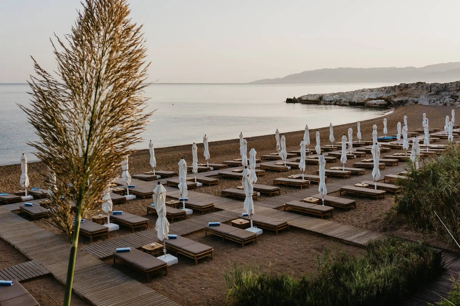 Luxury seaside beach club with closed white umbrellas and wooden loungers beside calm water