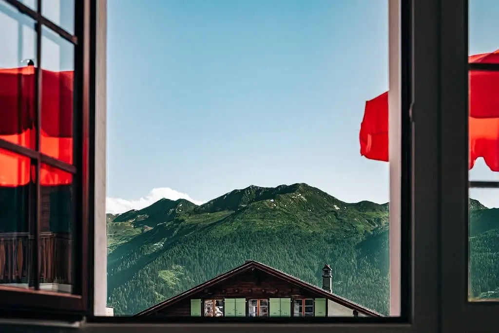 Red flags flutter in a chalet window as mountain peaks rise in the distance.