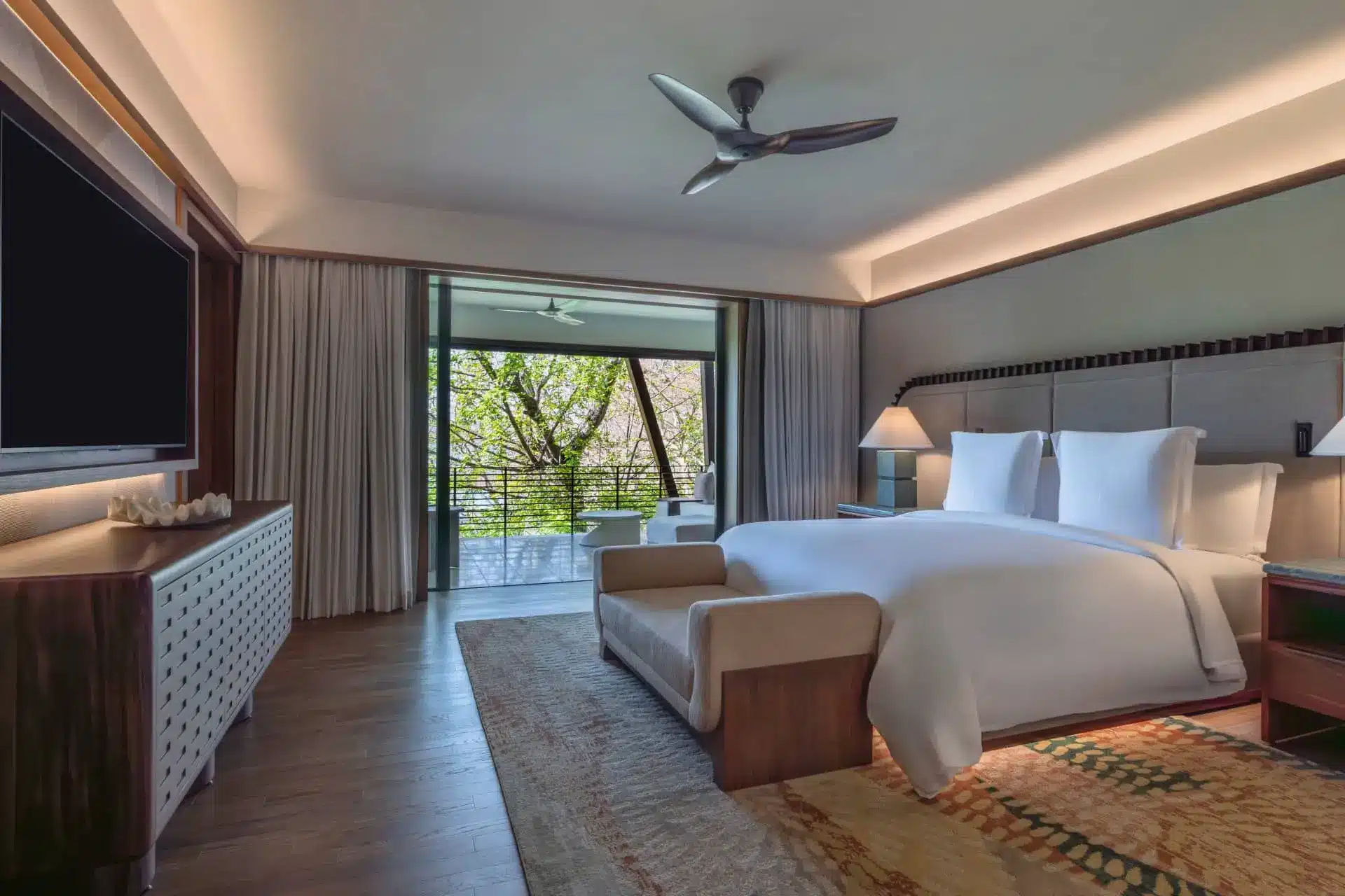 Luxury hotel bedroom with a king bed, balcony doors, and leafy views beyond