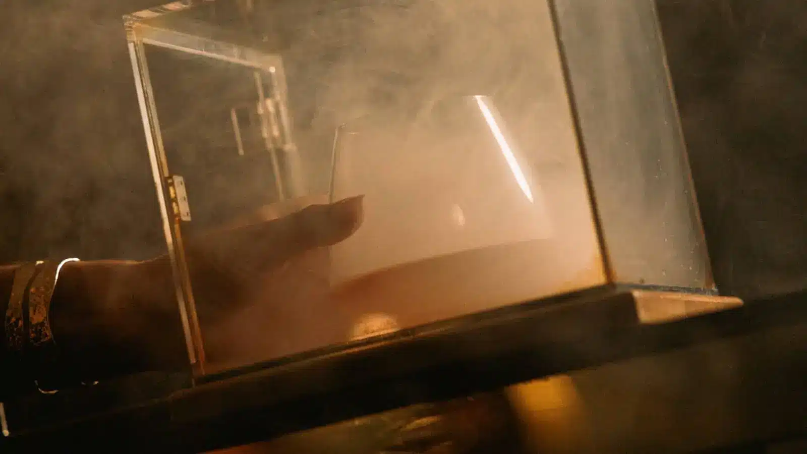Hand reaches toward an open translucent book-like prop in a smoky, dimly lit setting