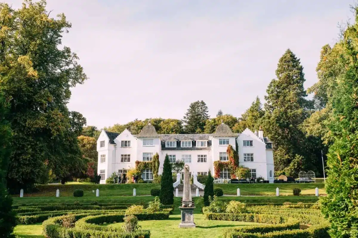 Grand white manor house overlooking manicured formal gardens and a central stone statue