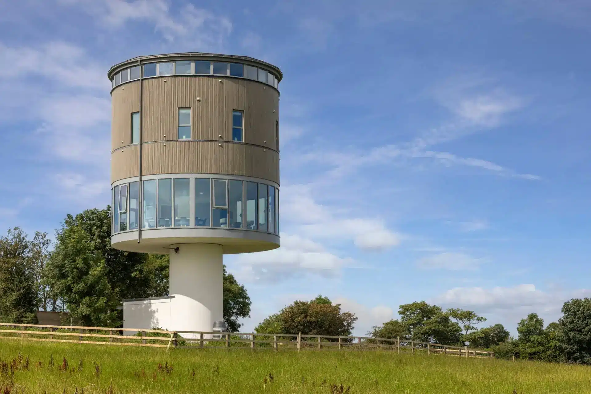 Cylindrical glass-and-wood tower rising above a grassy countryside meadow