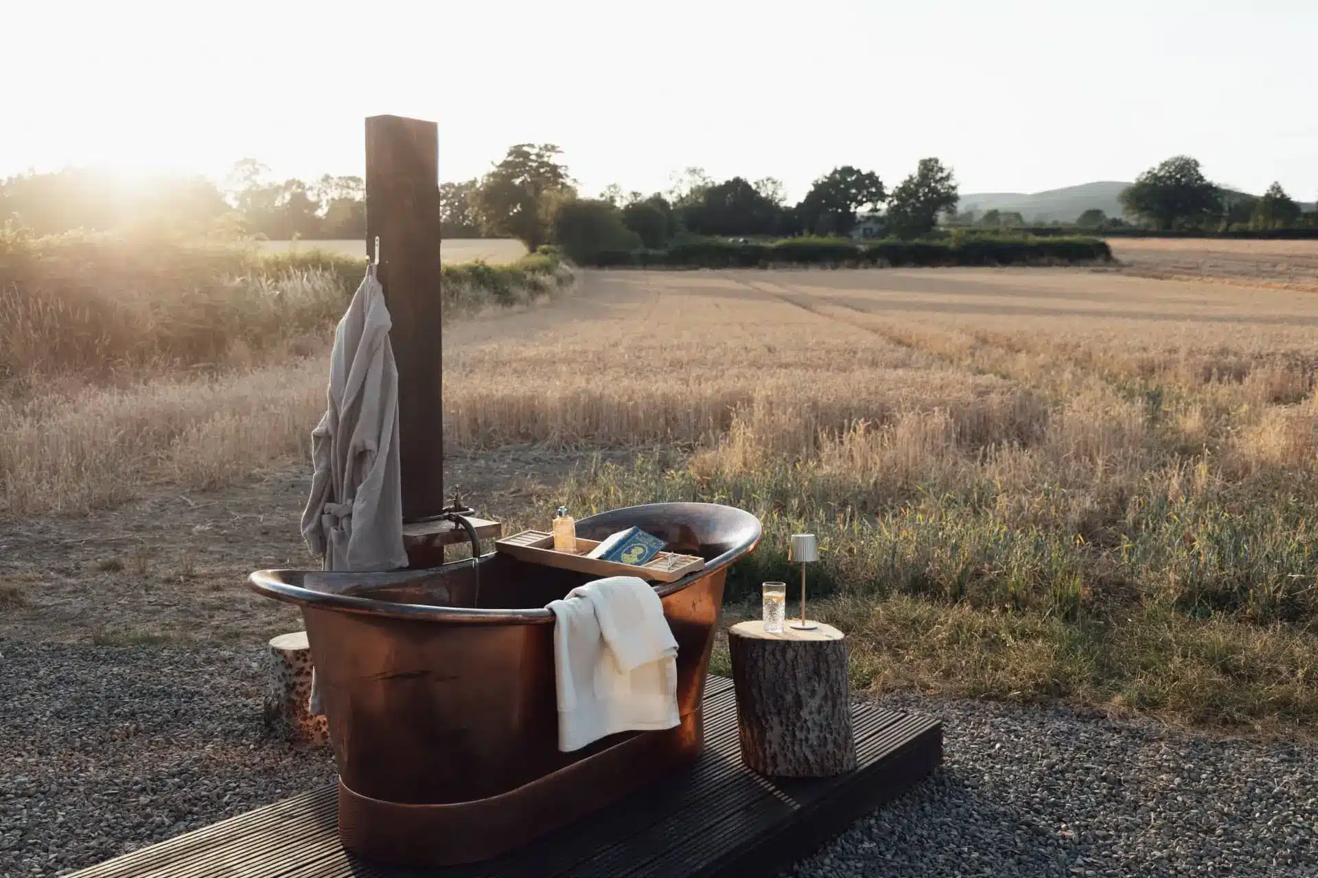 Rustic outdoor bathtub set in a sunlit field with towels and a wooden washstand