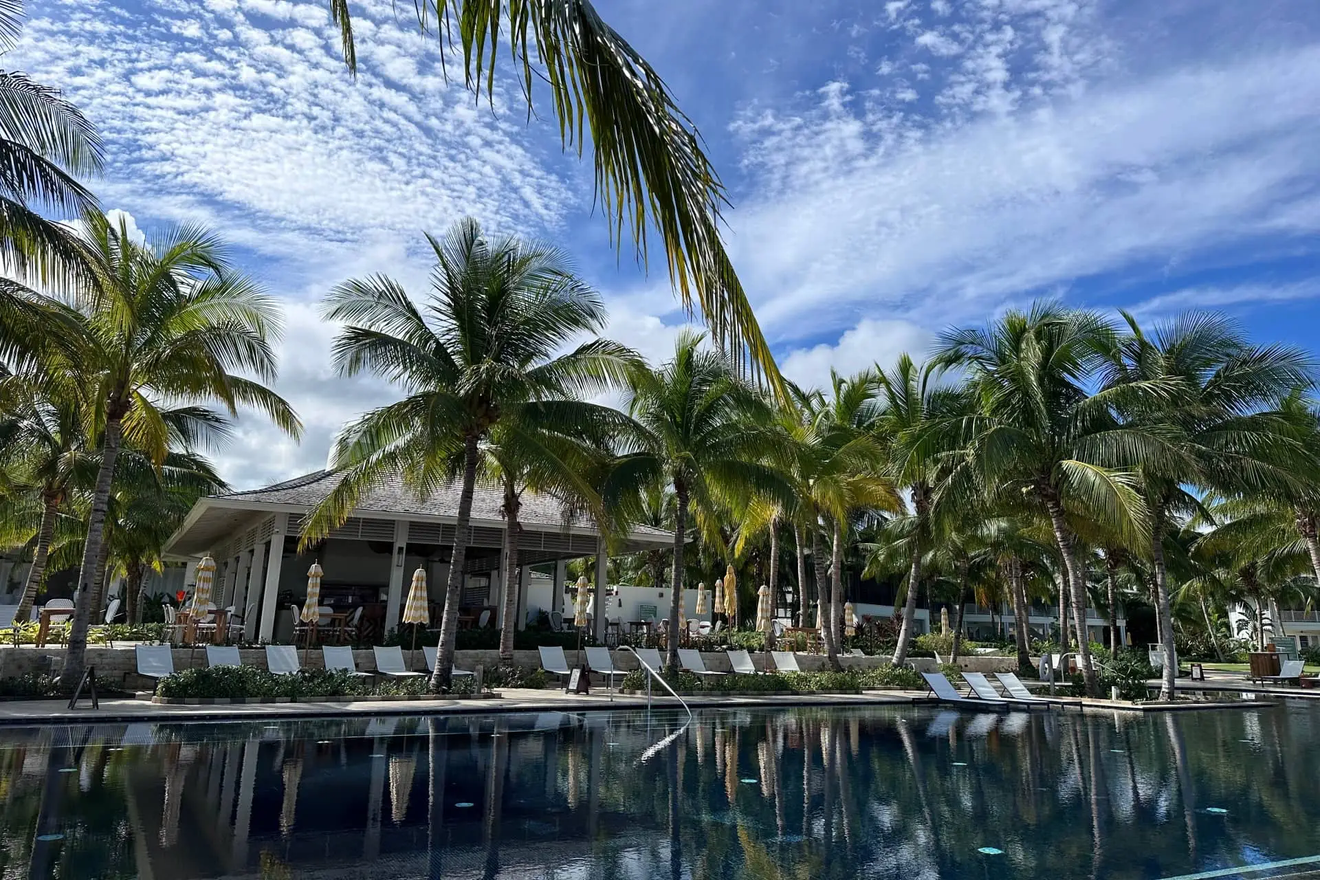 Palm-fringed resort pool with loungers and an open-air pavilion beneath a blue, cloud-dappled sky