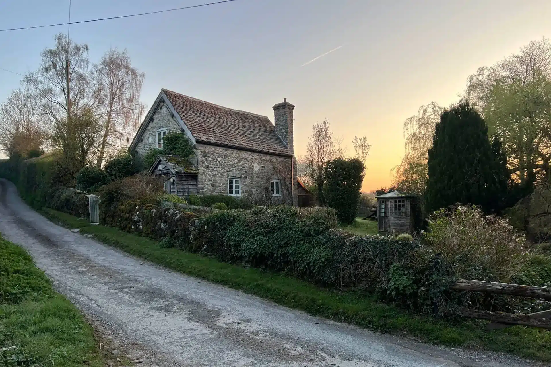 A quiet country lane curves past a stone cottage and clipped hedges at dusk.