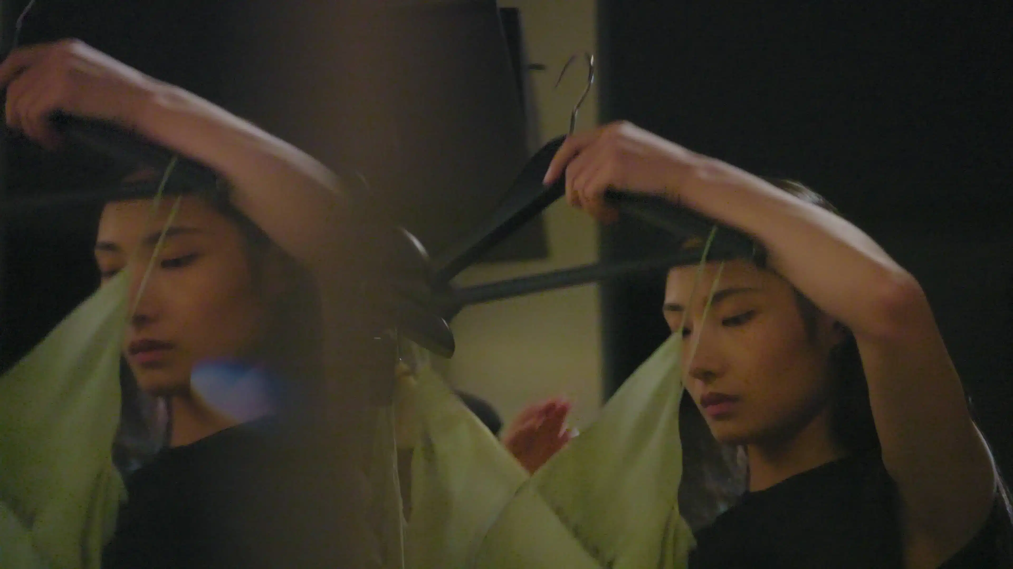 Immersive-theatre performer in black holds a long black prop beside her reflection, with green fabric behind her