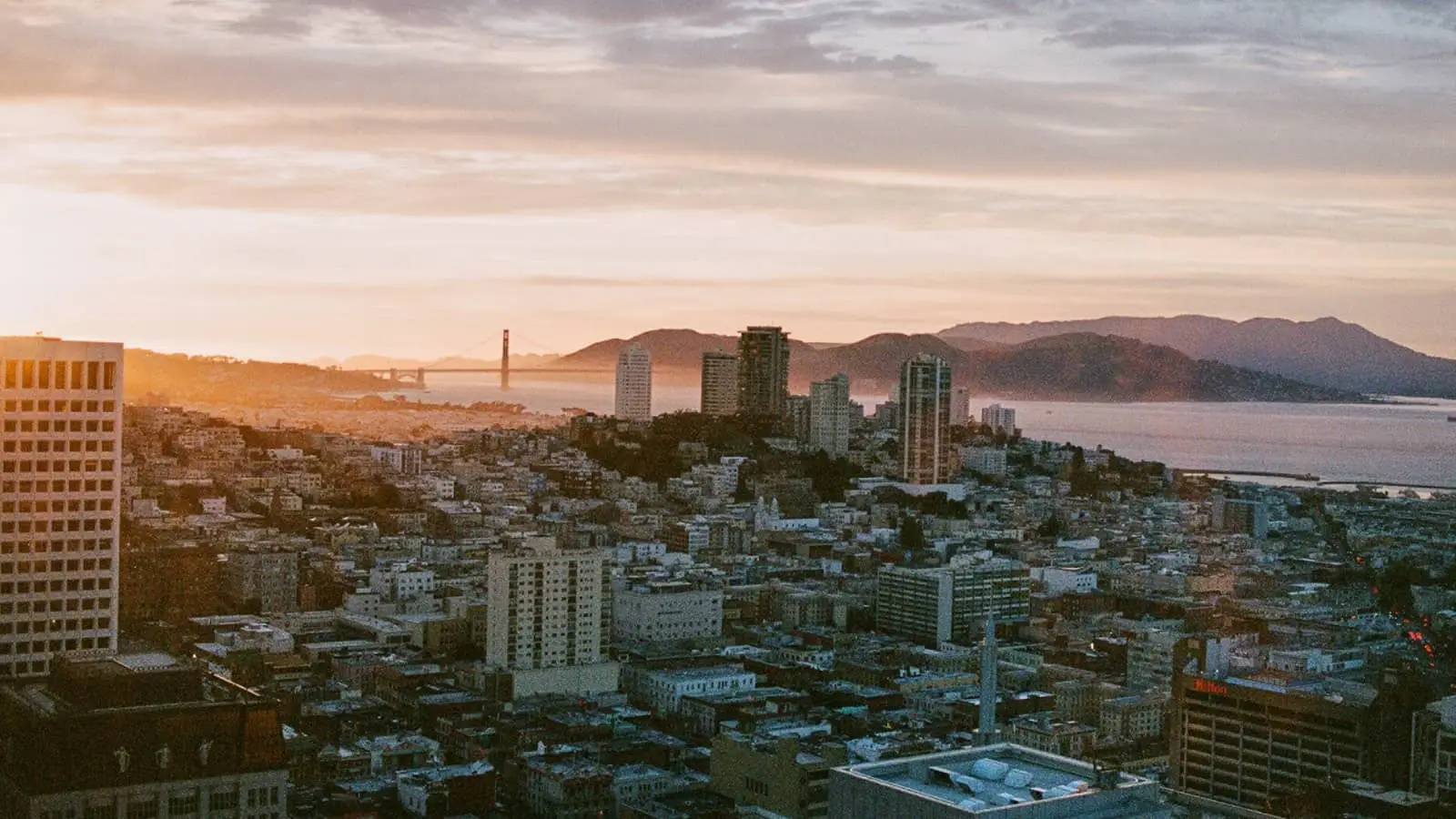 Sunset over a dense coastal city with high-rises, a broad bay, and mountains in the distance