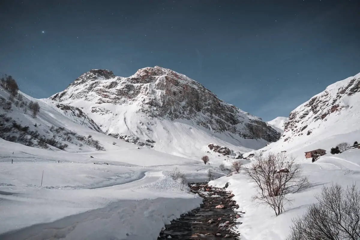 Snow-covered Face de Bellevarde rises above a ski run in Val d’Isère, with a small stream and bare trees in the foreground.
