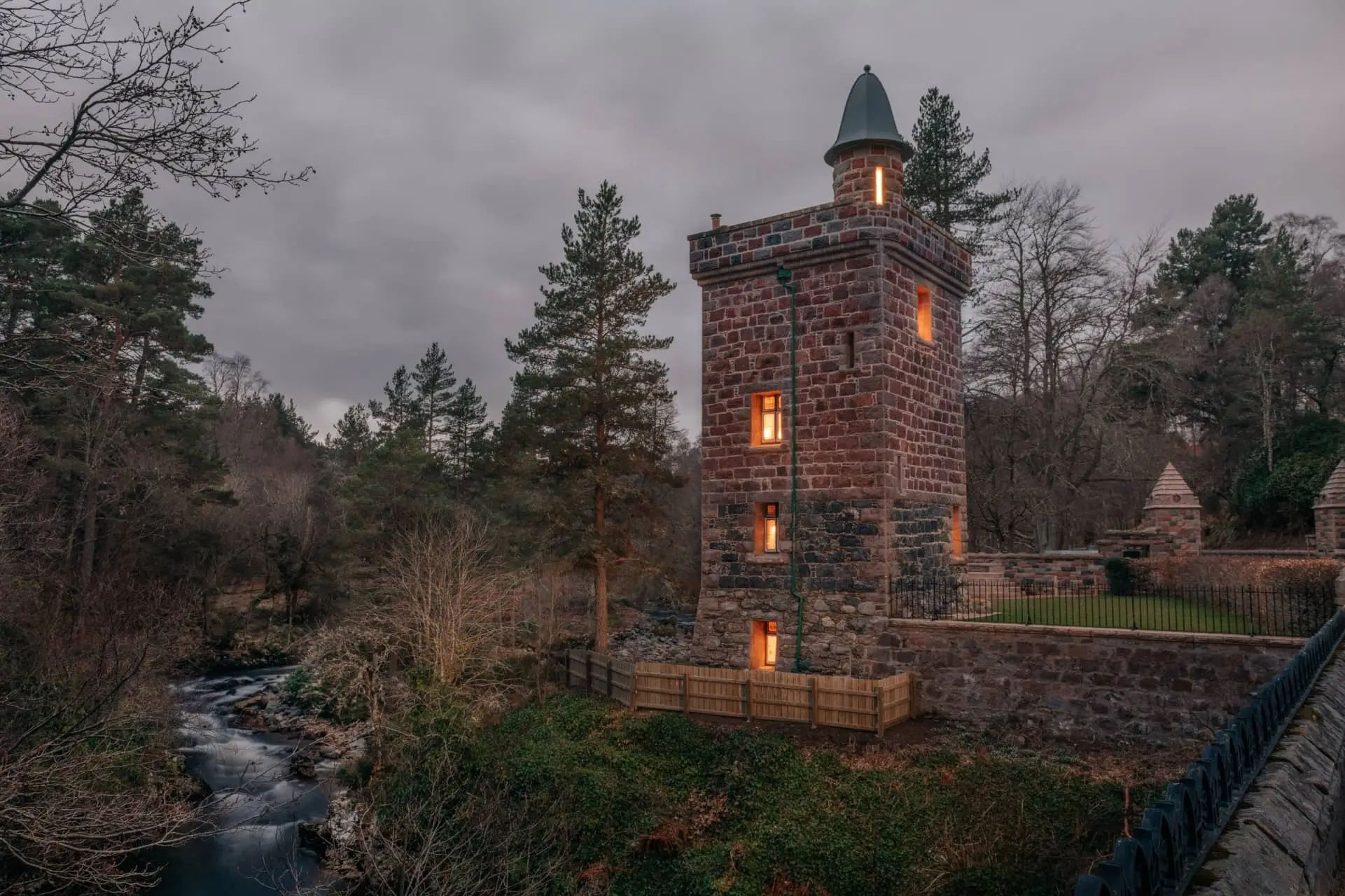 Historic brick tower with glowing windows beside a woodland stream on a cloudy evening
