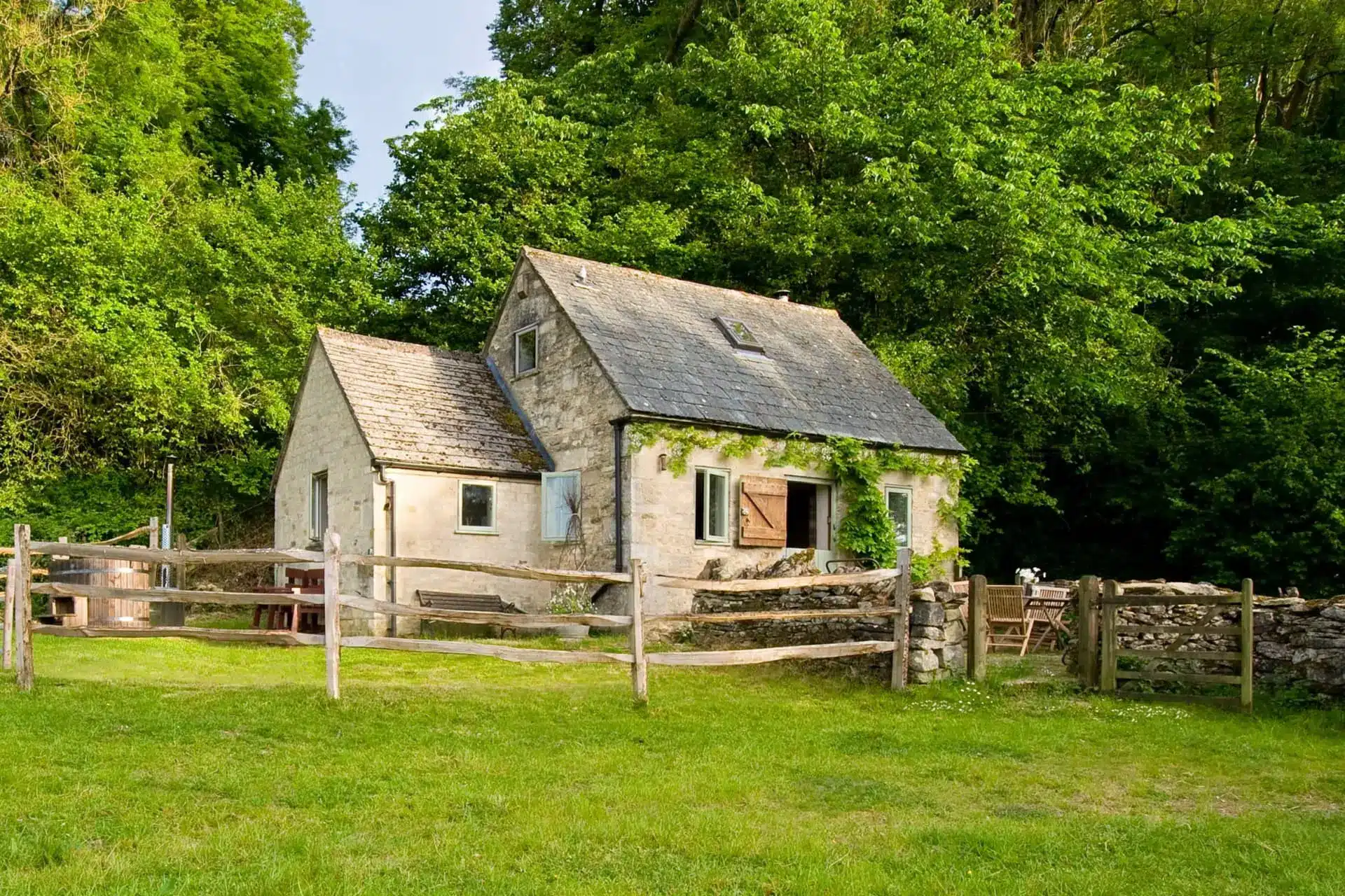 Stone cottage behind a rustic wooden fence, surrounded by green trees and a broad lawn