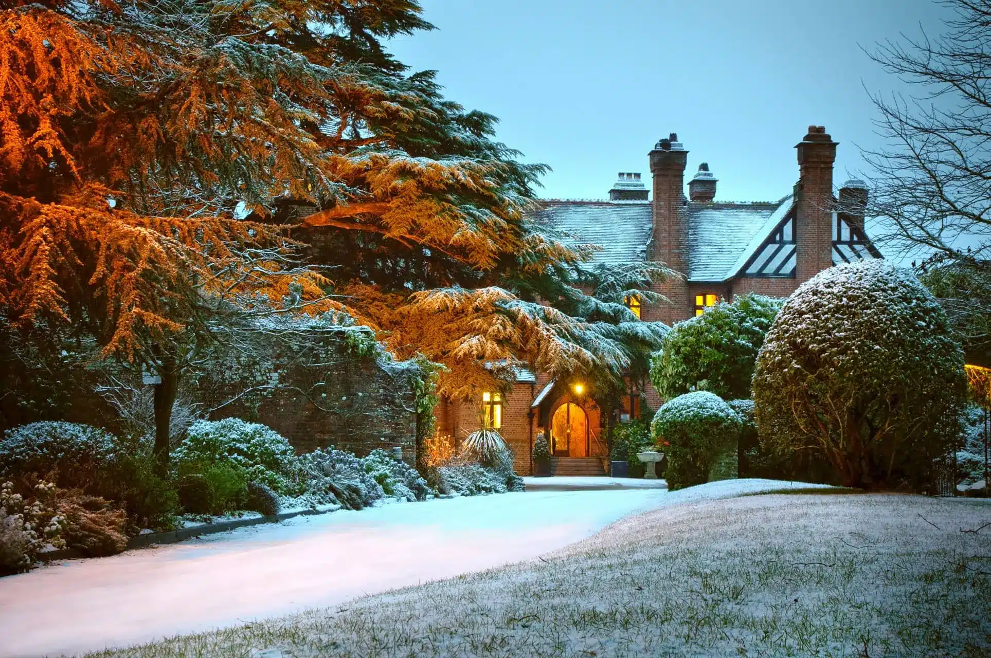 Snow-covered Careys Manor surrounded by evergreen trees, with a warmly lit entrance.