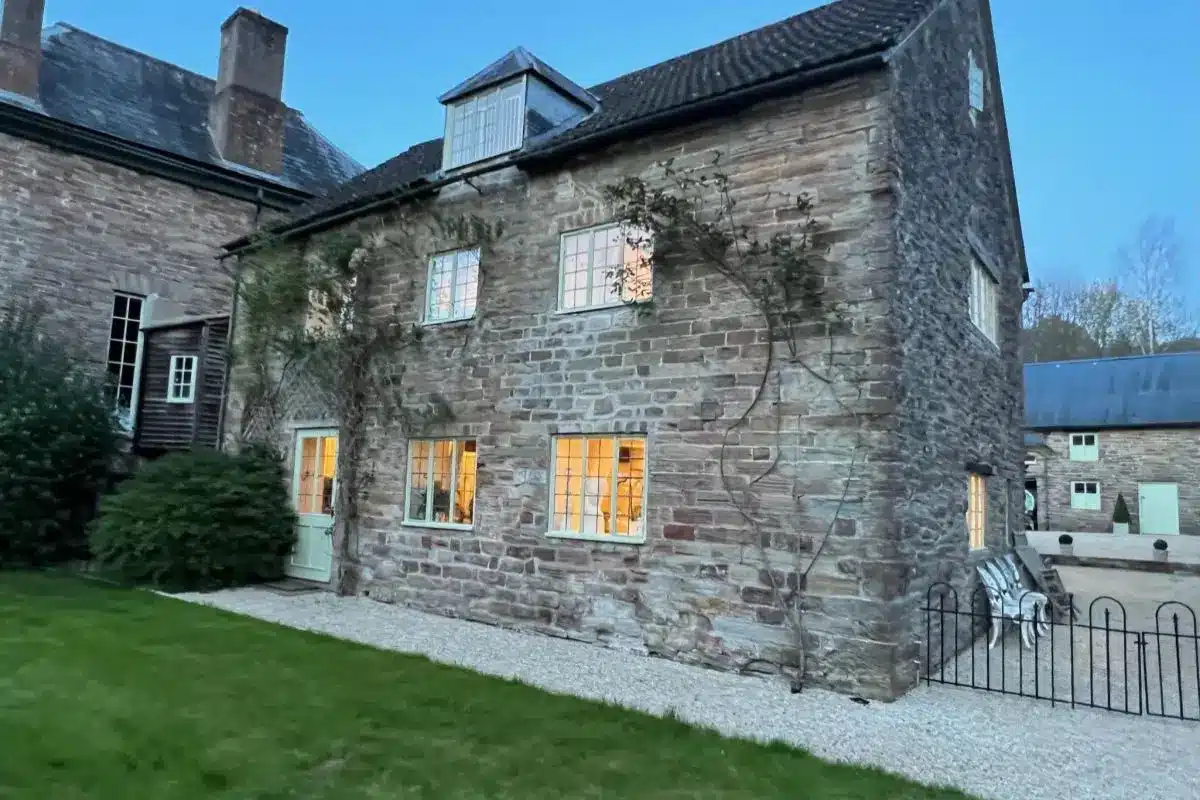 Stone manor house with warmly lit windows beside a green lawn and gravel path