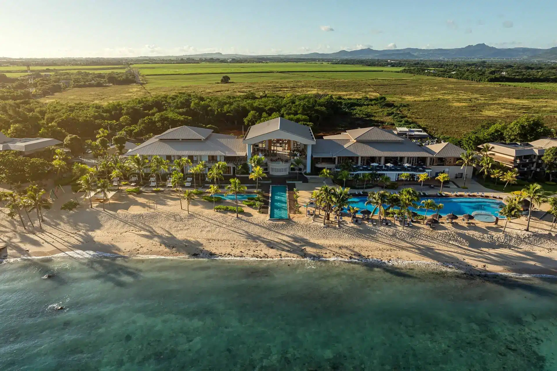 Aerial view of a luxury beachfront resort with pools, gardens, mountains, and turquoise ocean