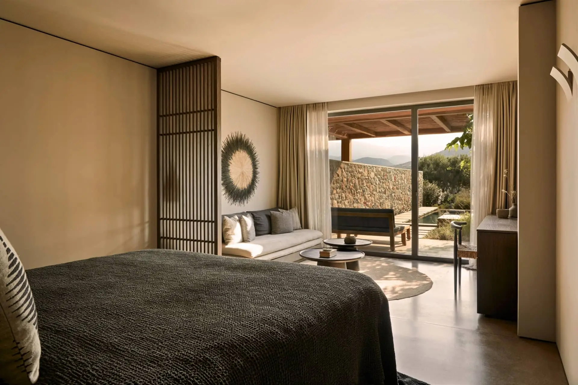 Luxury resort bedroom opening onto a sunlit terrace with a stone wall and mountain view