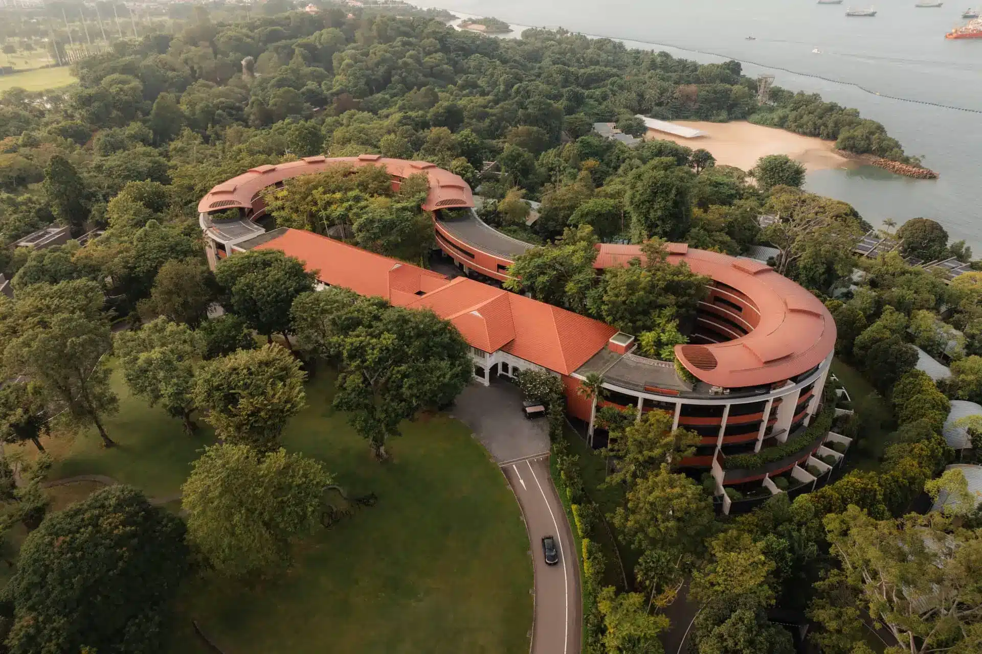 Aerial view of Capella Singapore’s red-roofed buildings amid lush grounds on Sentosa Island beside the sea.