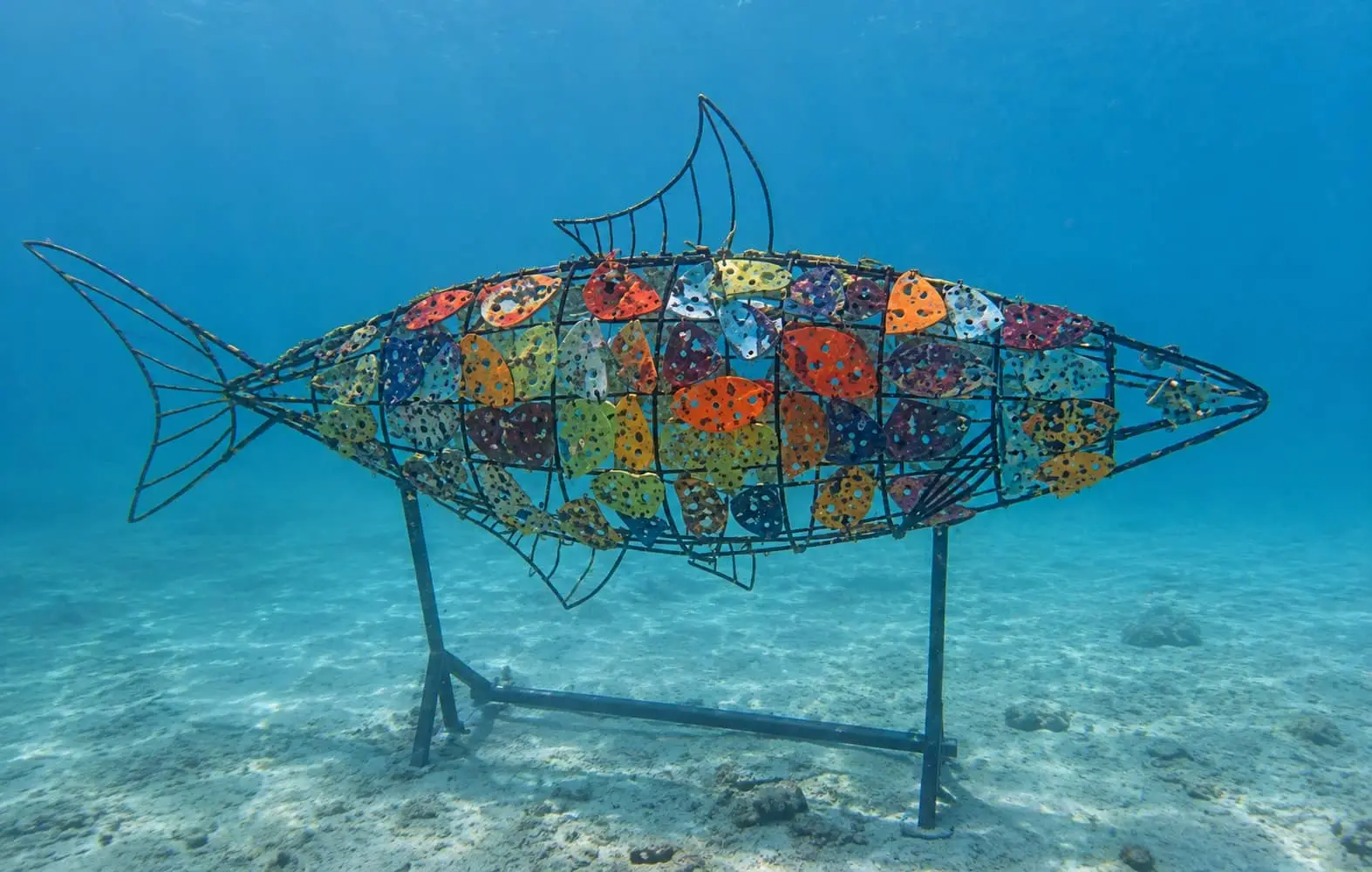 Colorful mosaic fish sculpture resting on a metal frame beneath clear blue seawater