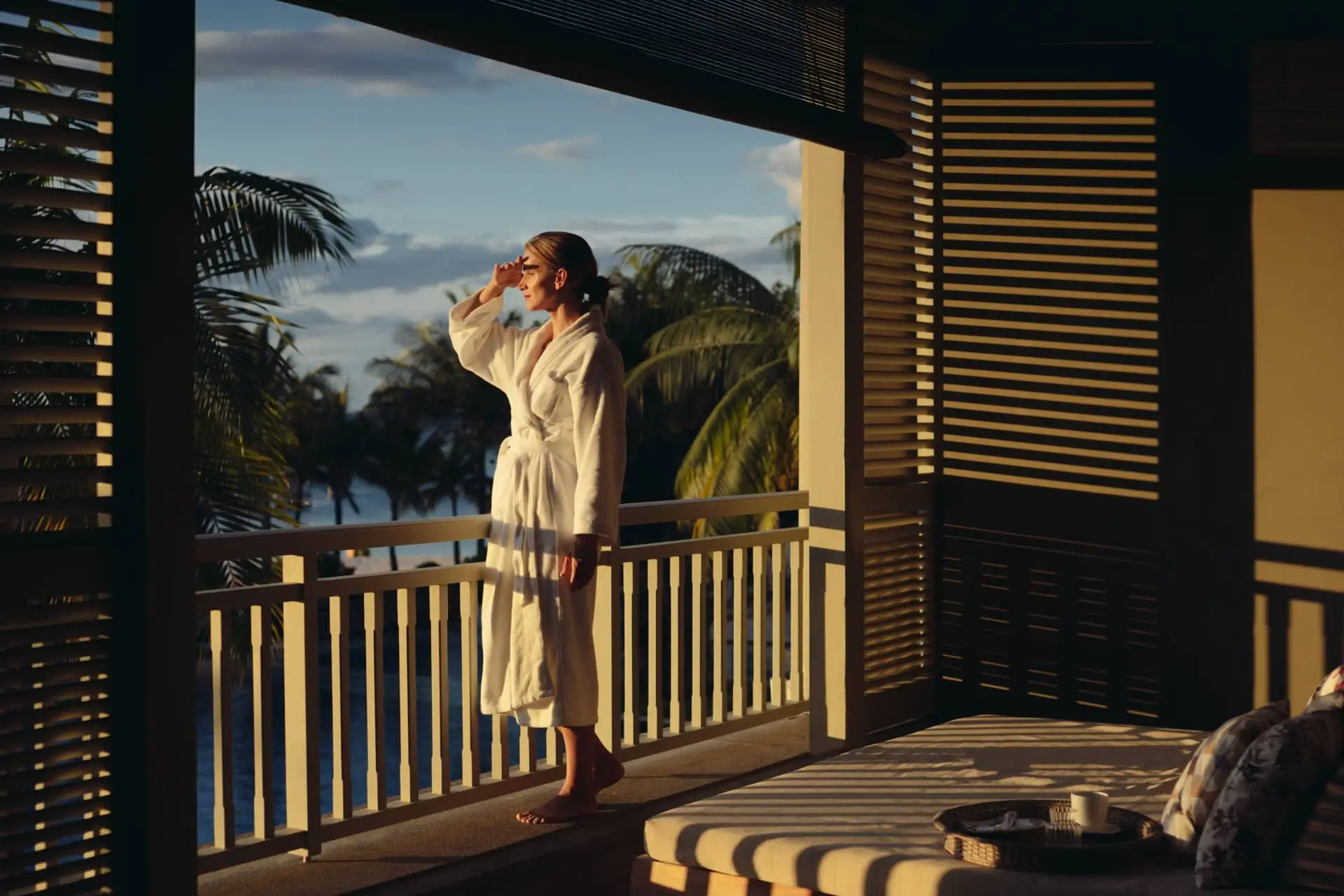 Woman enjoying a drink on a sunlit balcony overlooking a tropical coastline