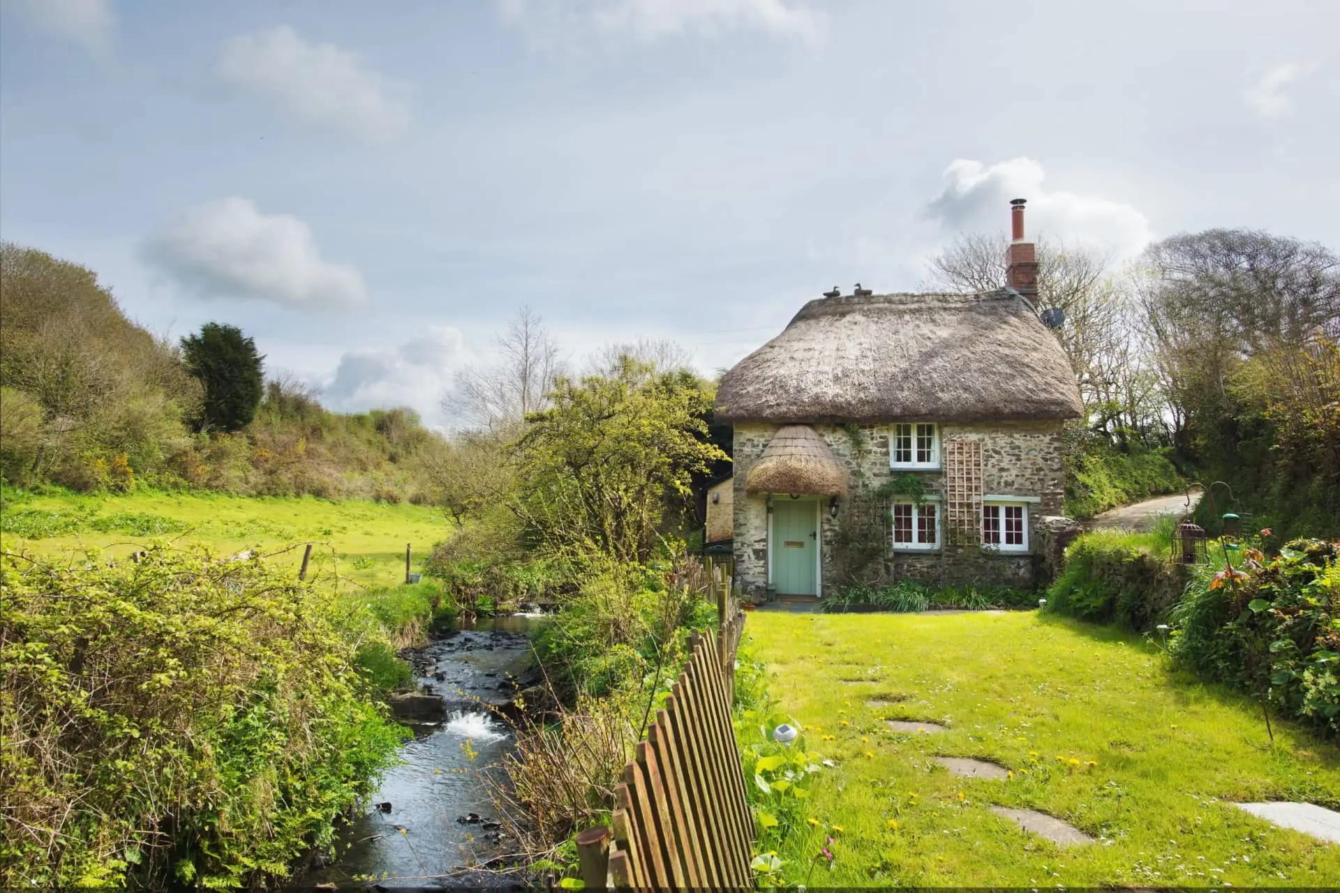 Thatched-roof cottage beside a narrow stream in a lush countryside garden