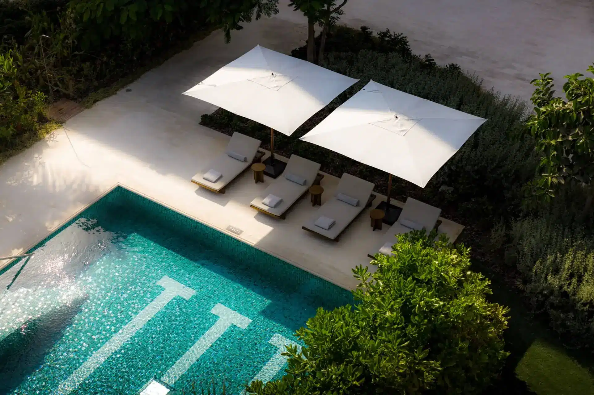 A turquoise resort pool beside white umbrellas and sun loungers on a landscaped terrace
