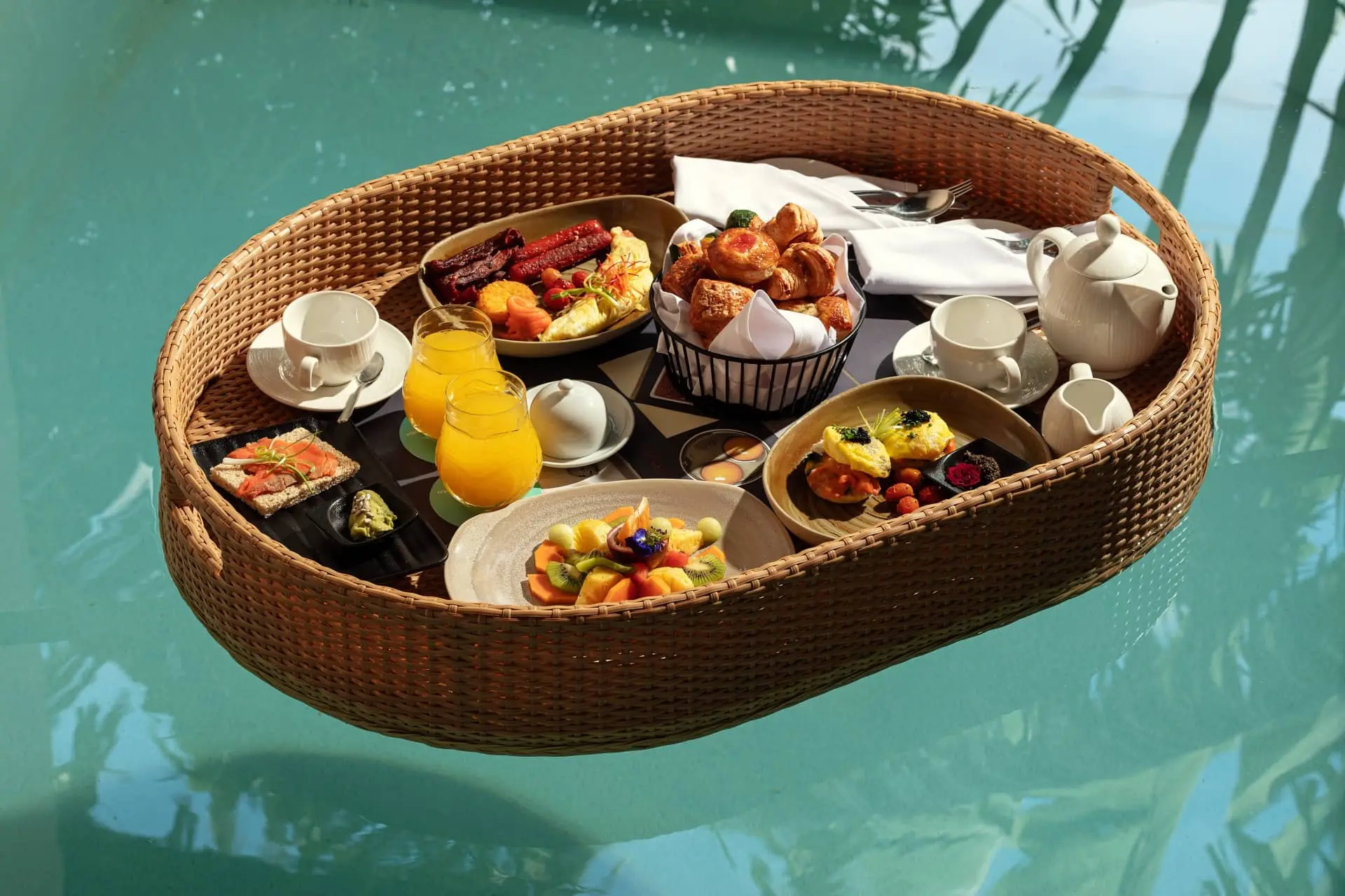 Floating wicker breakfast basket filled with pastries, fruit, coffee, and juice in a turquoise pool