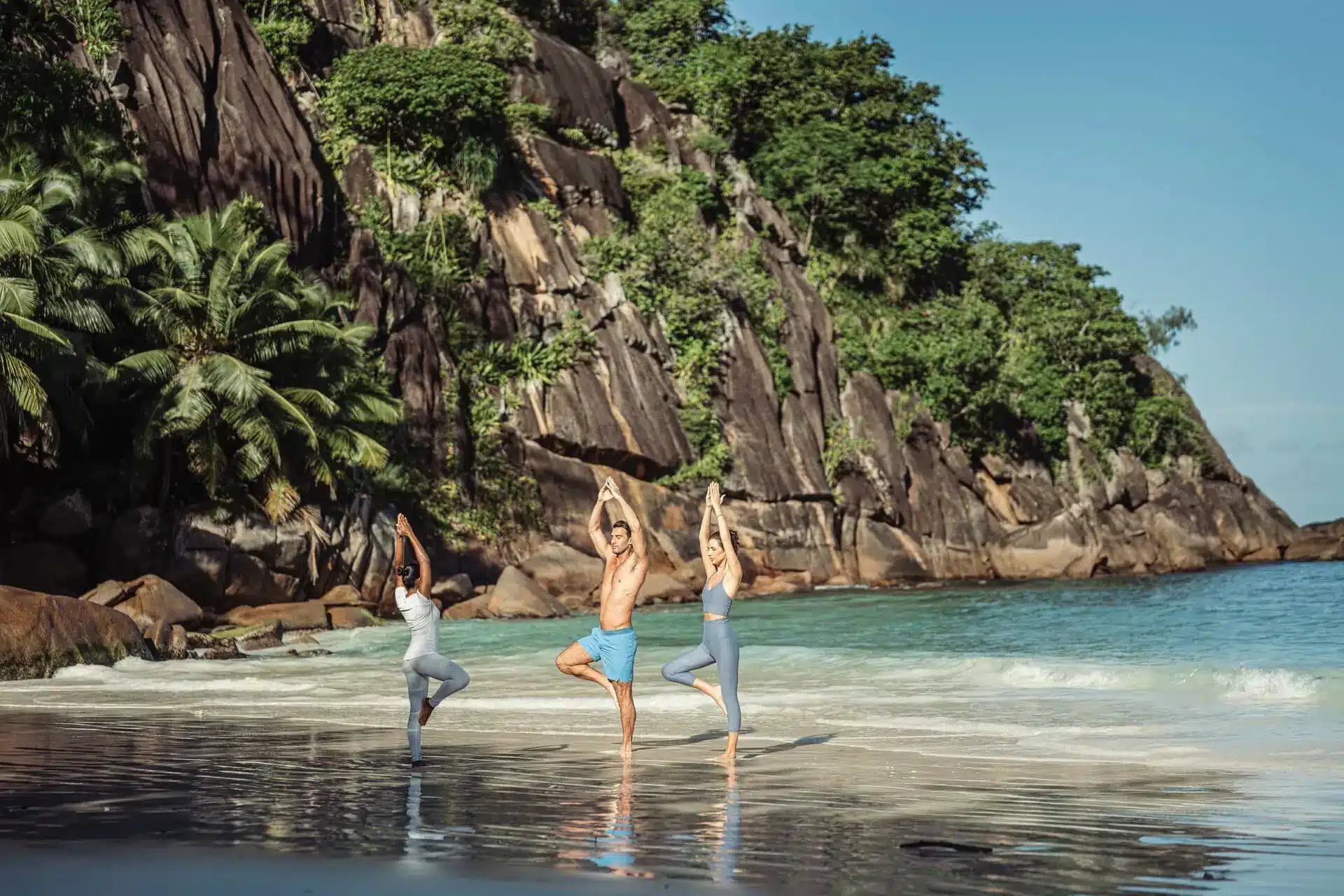 Three travelers practice yoga in shallow water on a tropical beach beneath a rocky cliff.
