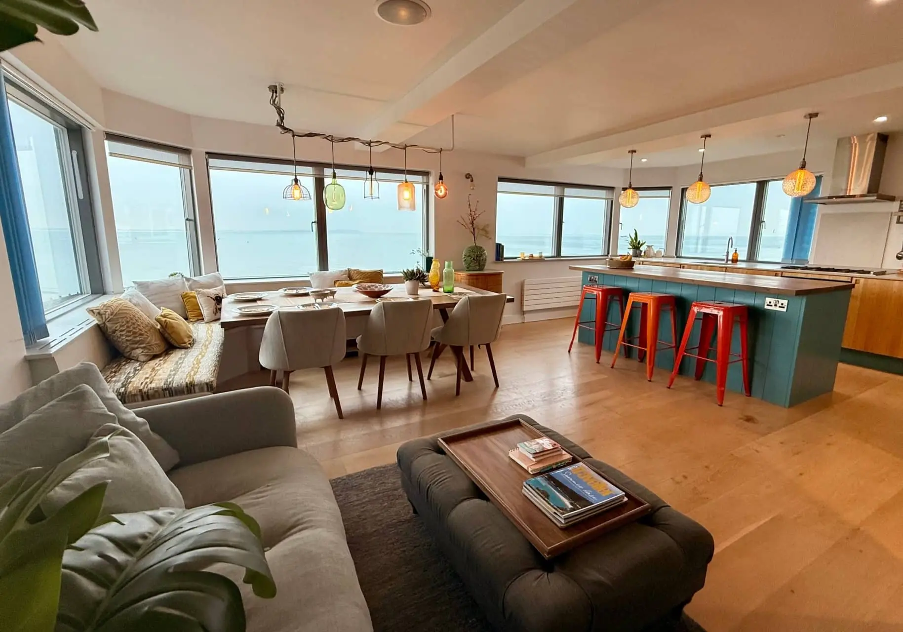 Open-plan living and dining area with a grey sofa, dining table and kitchen island beside large windows.