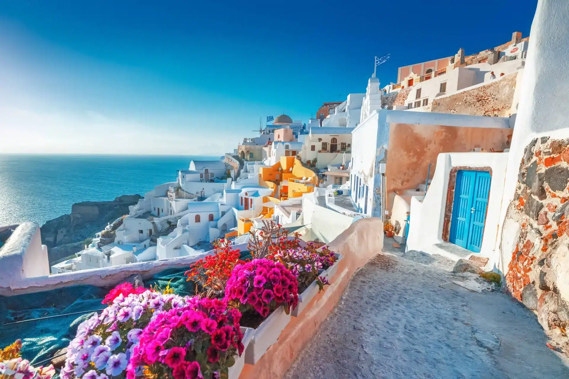 Whitewashed Mediterranean buildings overlook the sea, with bright pink flowers along a terrace.