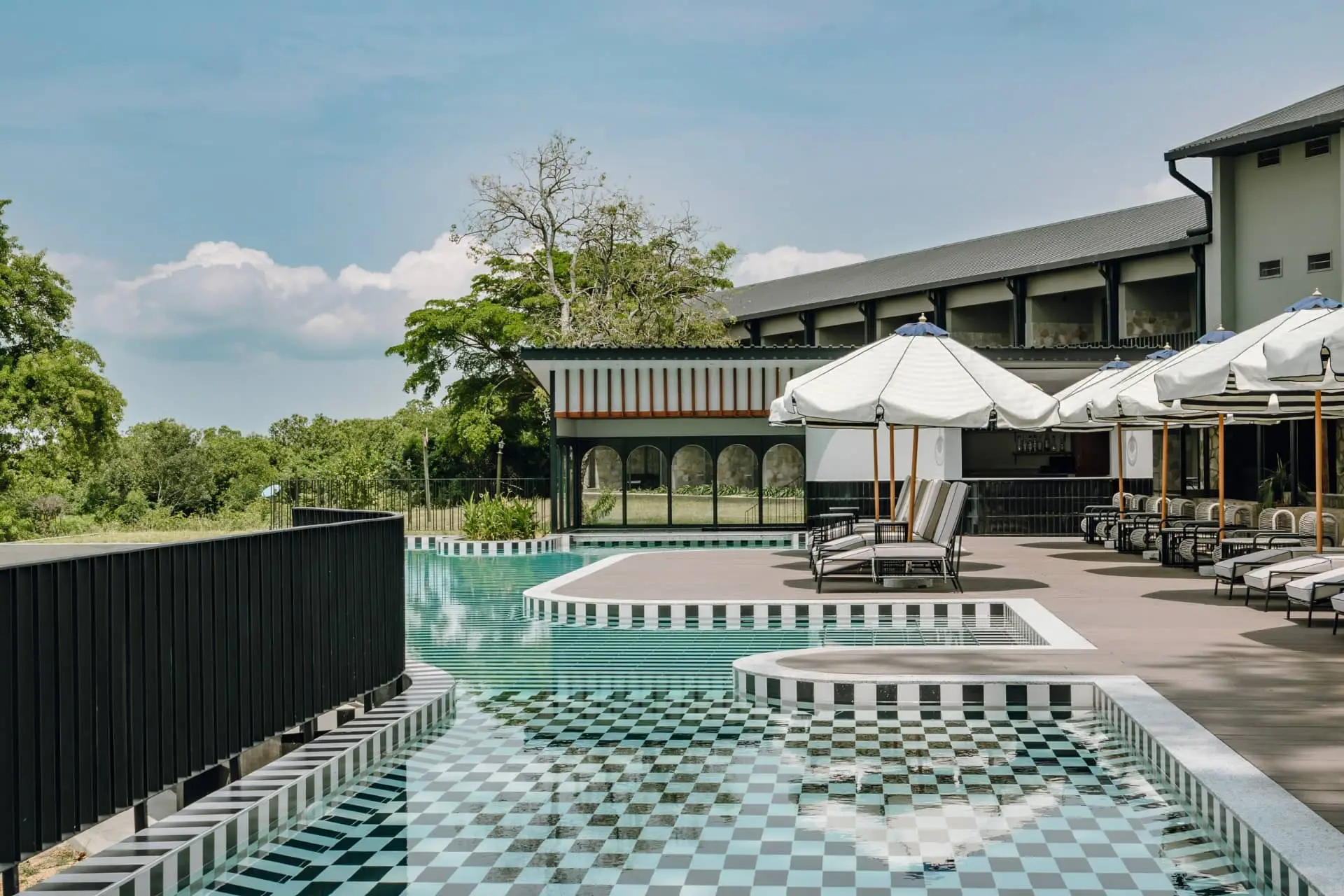 Quiet modern resort courtyard with an empty turquoise pool, checkerboard tiles, loungers, and white parasols