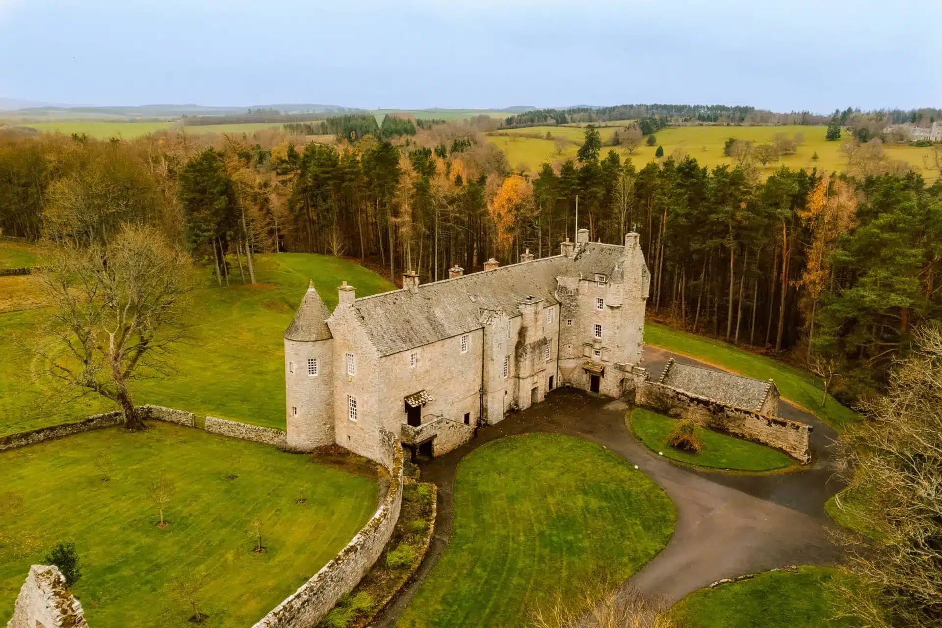 Aerial view of a stone castle with a circular driveway, green lawns and woodland beyond