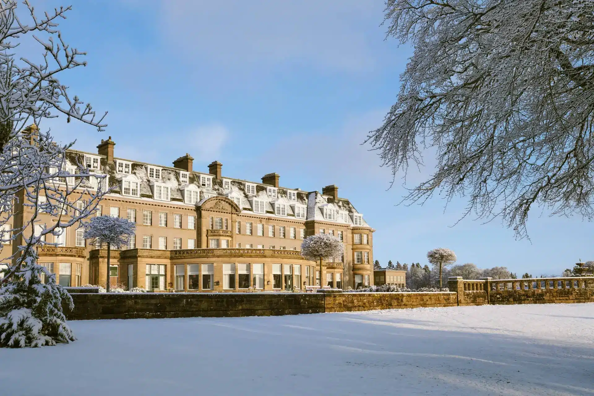 Gleneagles hotel in winter, with snow-covered grounds and bare trees beneath a clear blue sky.
