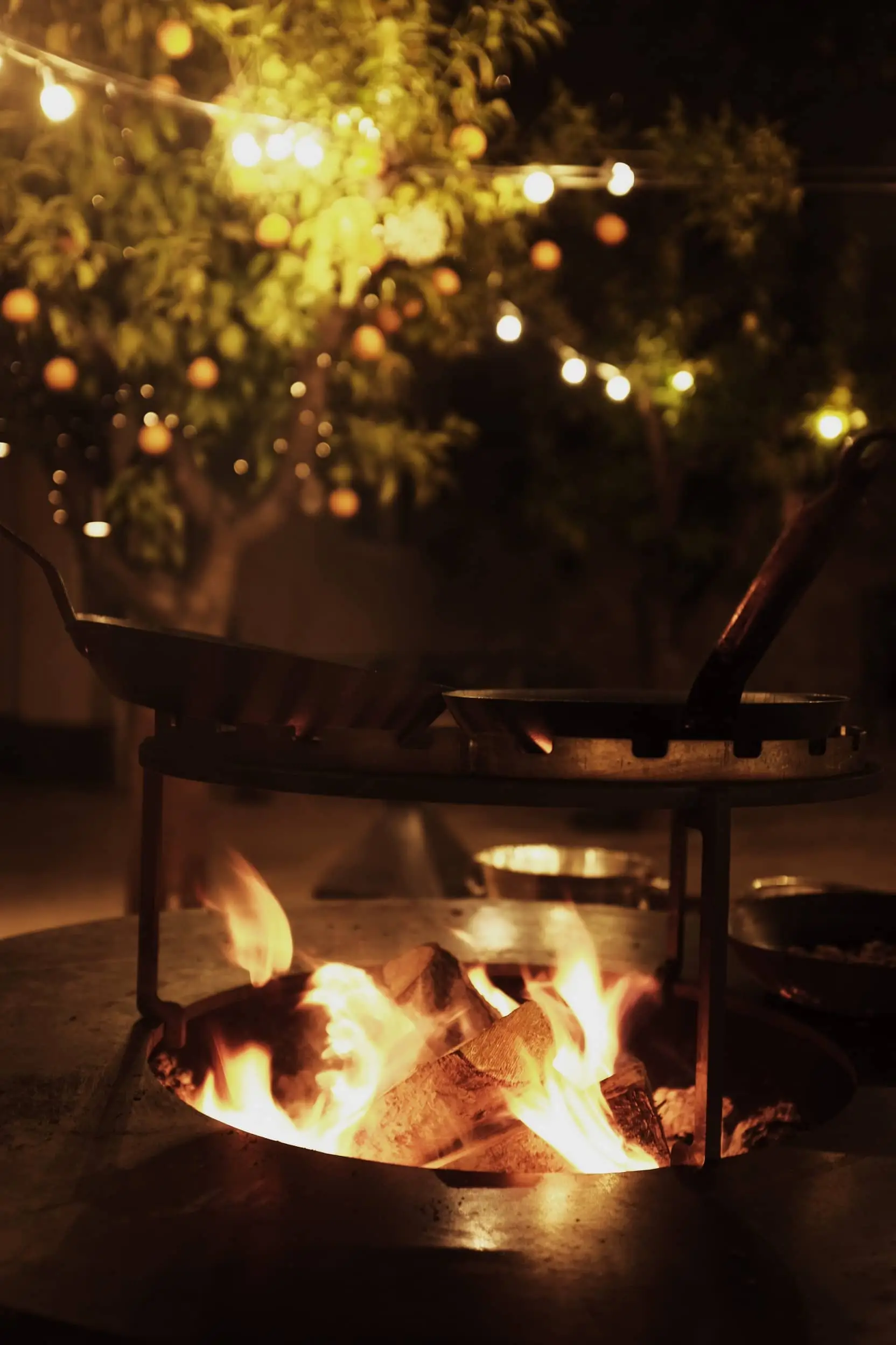 Fire burns in a hearth beneath a dark metal canopy, with warm lights glowing in the background.
