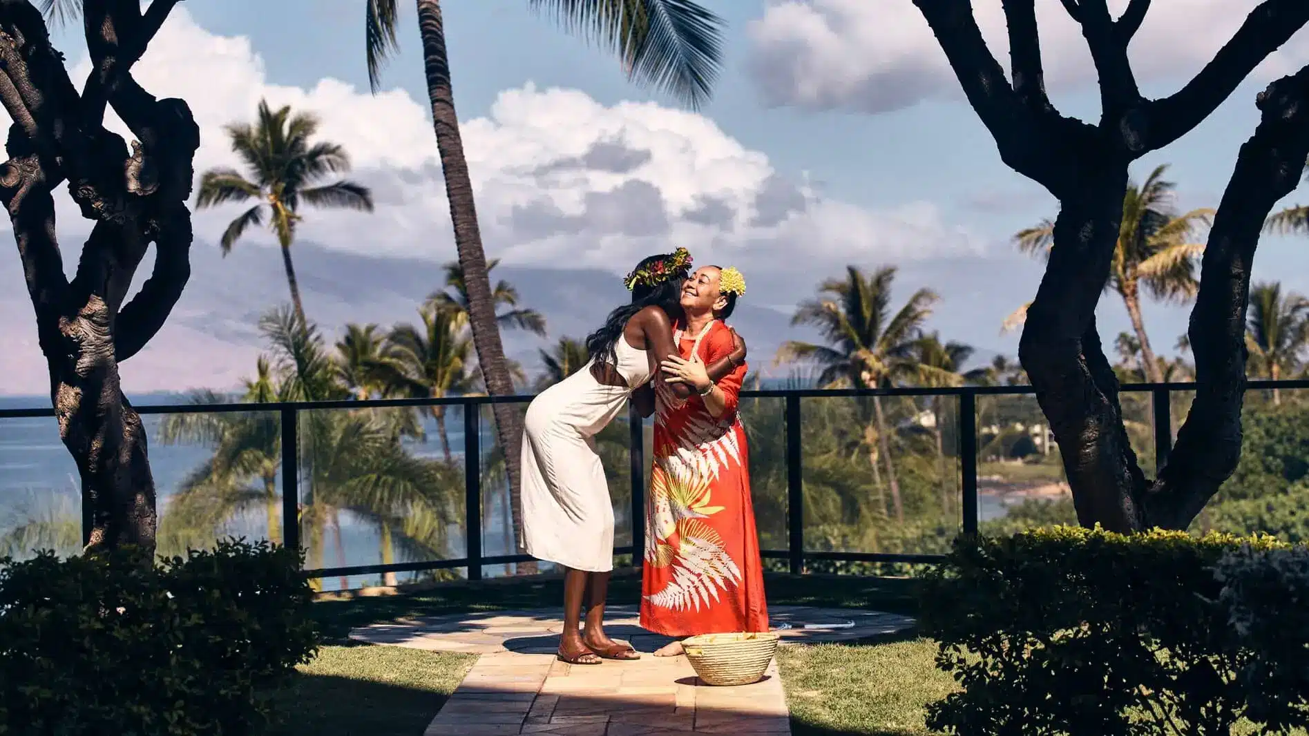 A couple in traditional Hawaiian attire embrace beside a garden path, with palms and the resort grounds behind them.