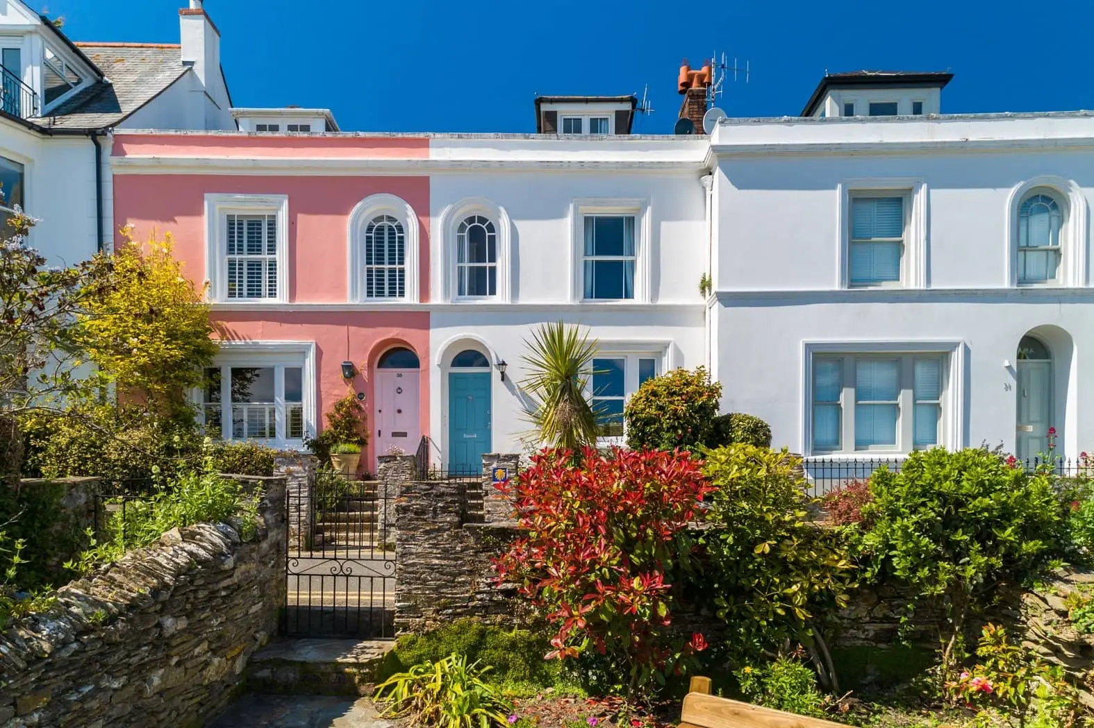 The Moorings, a pale waterfront house in St Mawes, with a stone wall and flowering shrubs in front.