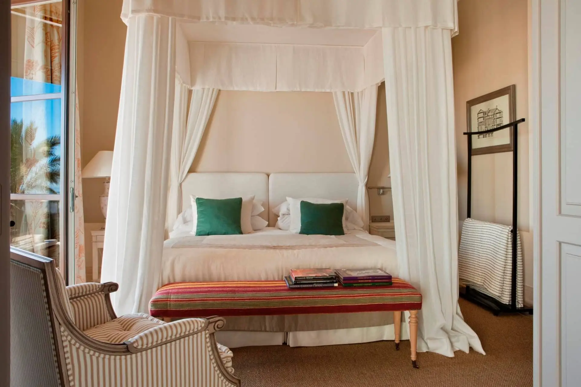 Canopy bed with white drapes, teal cushions and a red bench in a bright hotel suite