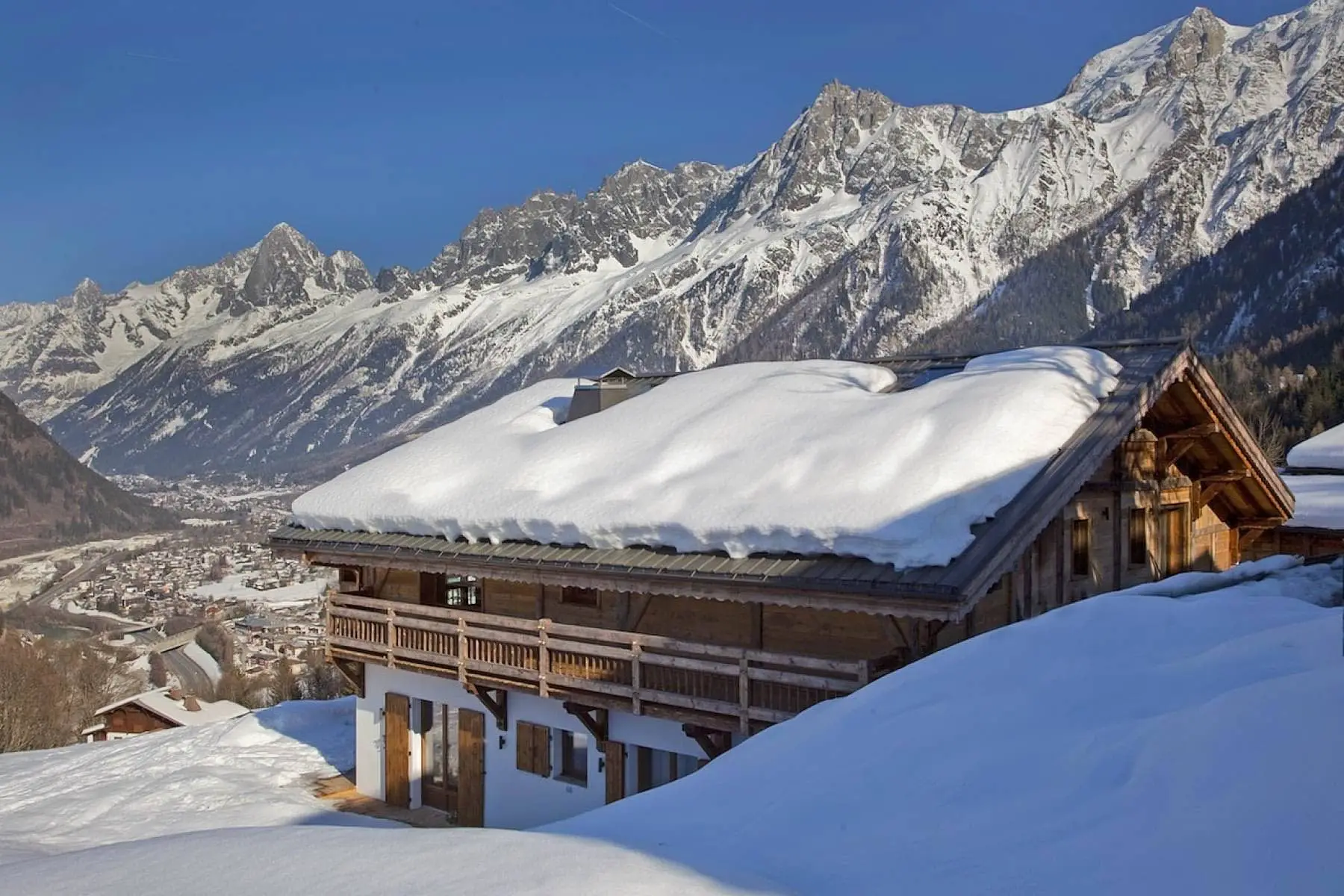 Snow-covered Alpine chalet beneath the mountains, with a broad roof and balconies overlooking a winter landscape.