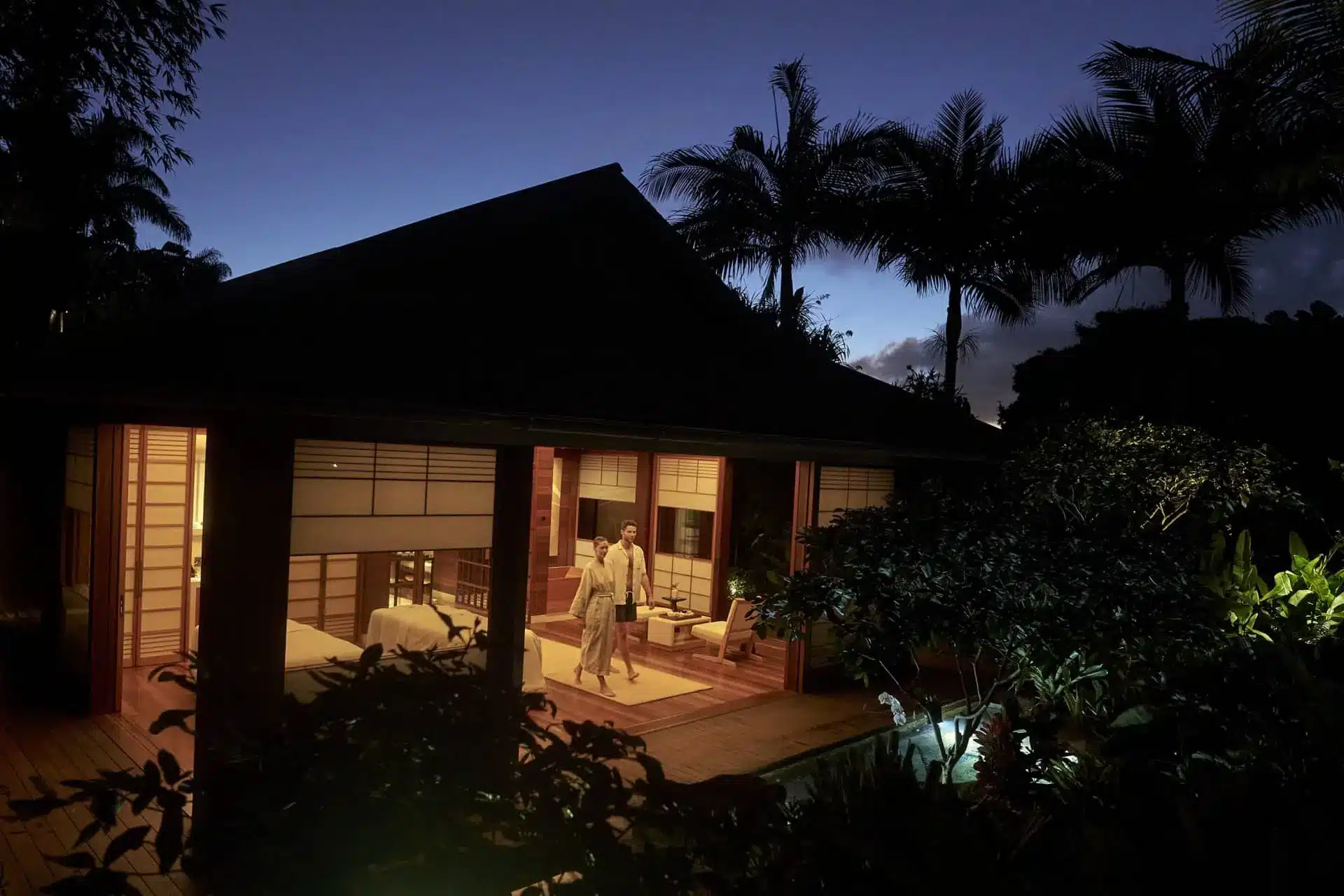 A couple stands inside a warmly lit luxury villa as dusk falls over the tropical garden.