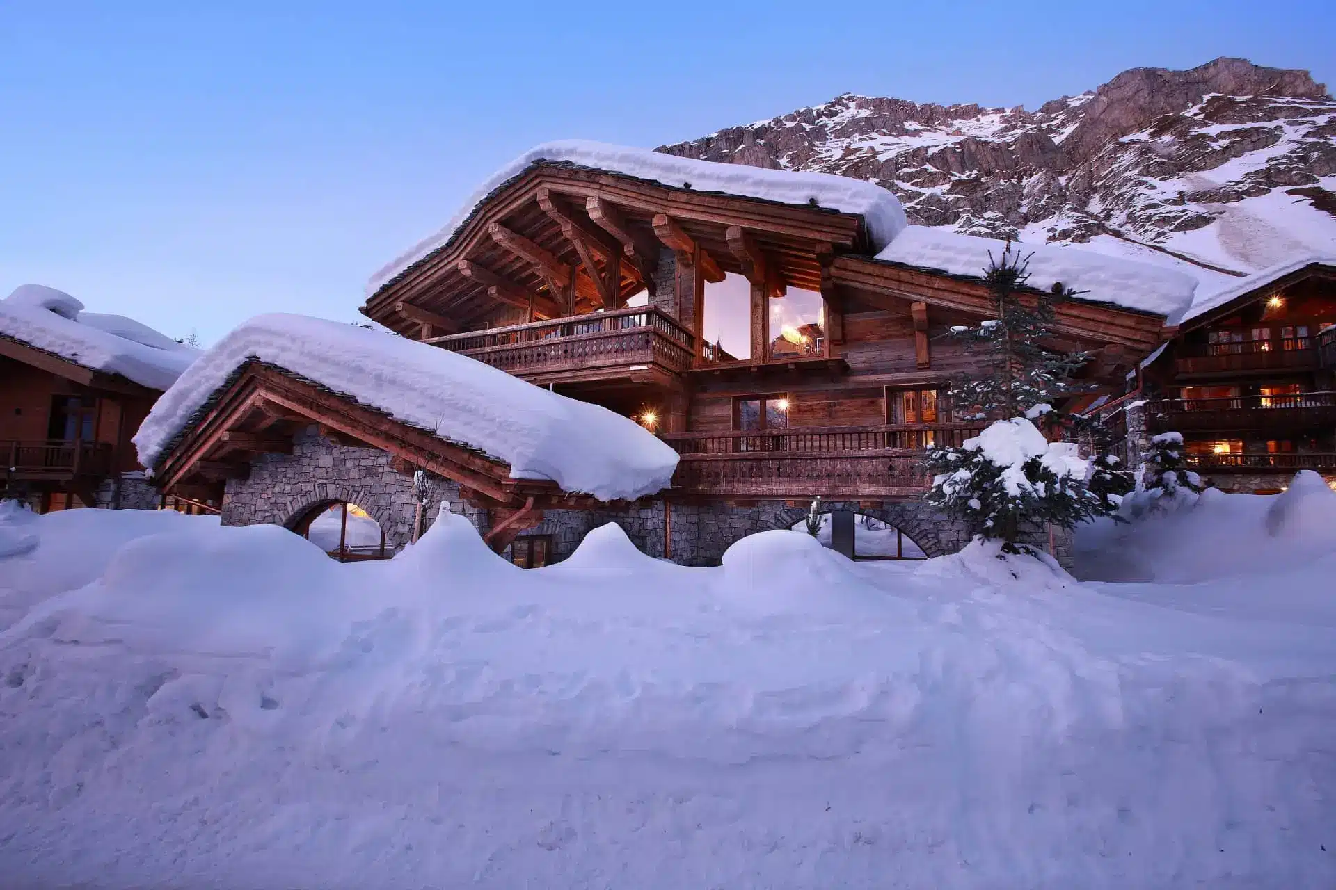 Snow-covered chalet with timber balconies and warm-lit windows, set against a mountain slope.