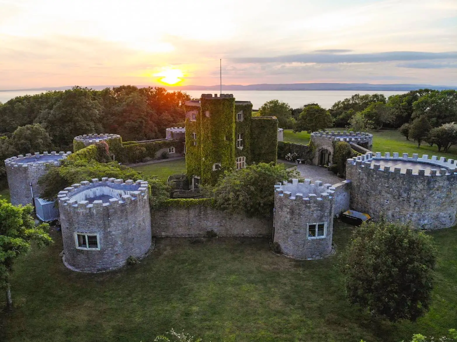 Aerial view of C17th Castle’s central keep and round turrets, surrounded by lawns and trees at sunset.