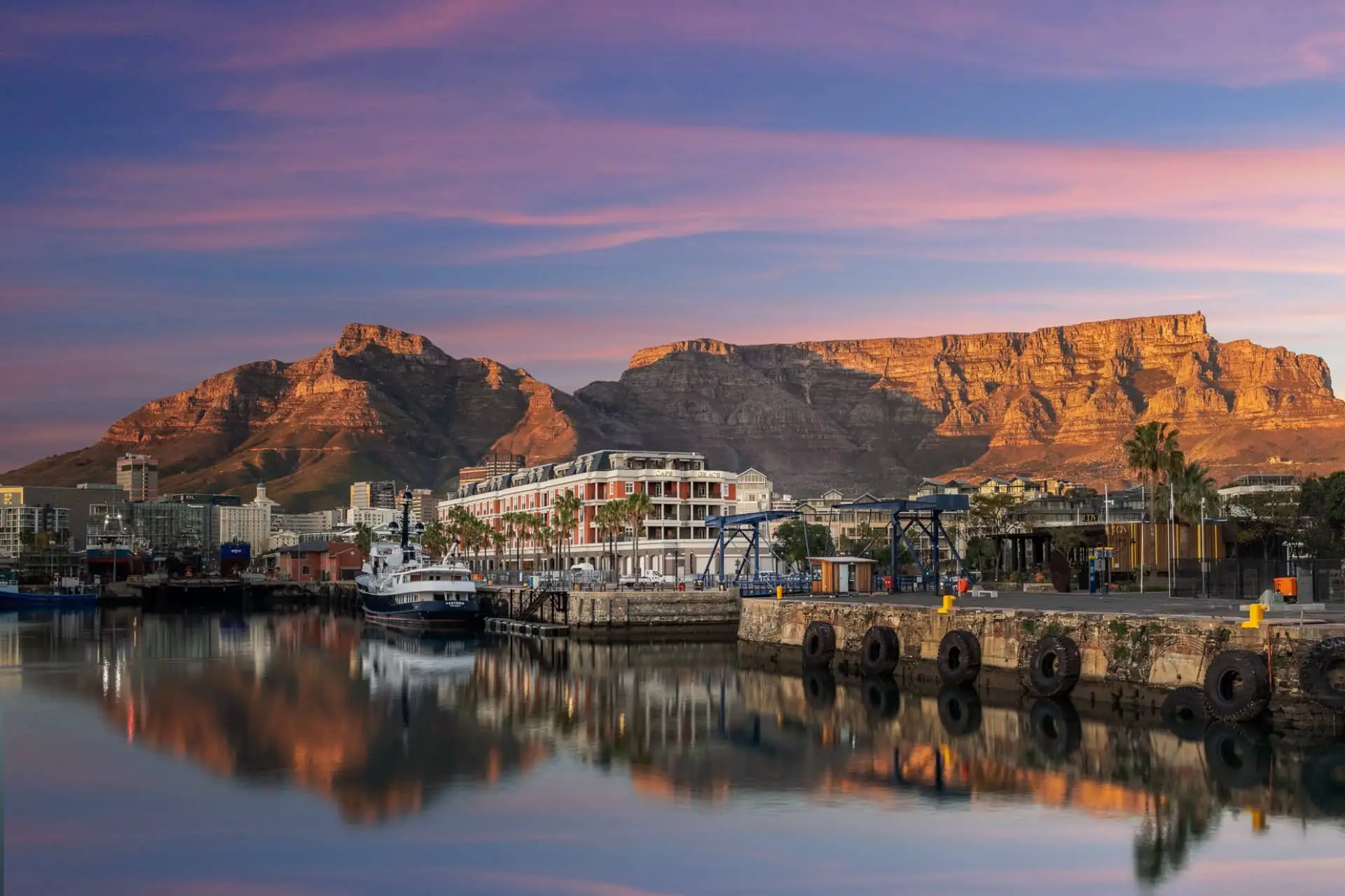 Cape Grace hotel and waterfront buildings reflected in the marina, with Table Mountain glowing behind them at sunset.