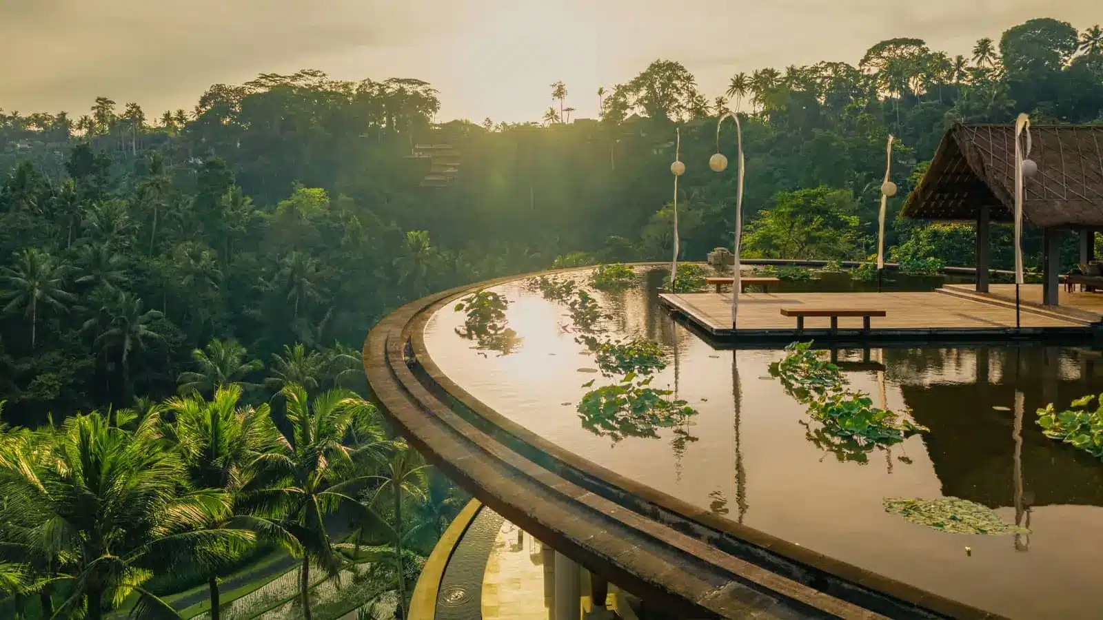 A curved infinity pool at Four Seasons Resort Bali at Sayan overlooks a lush, tree-covered valley at sunset.